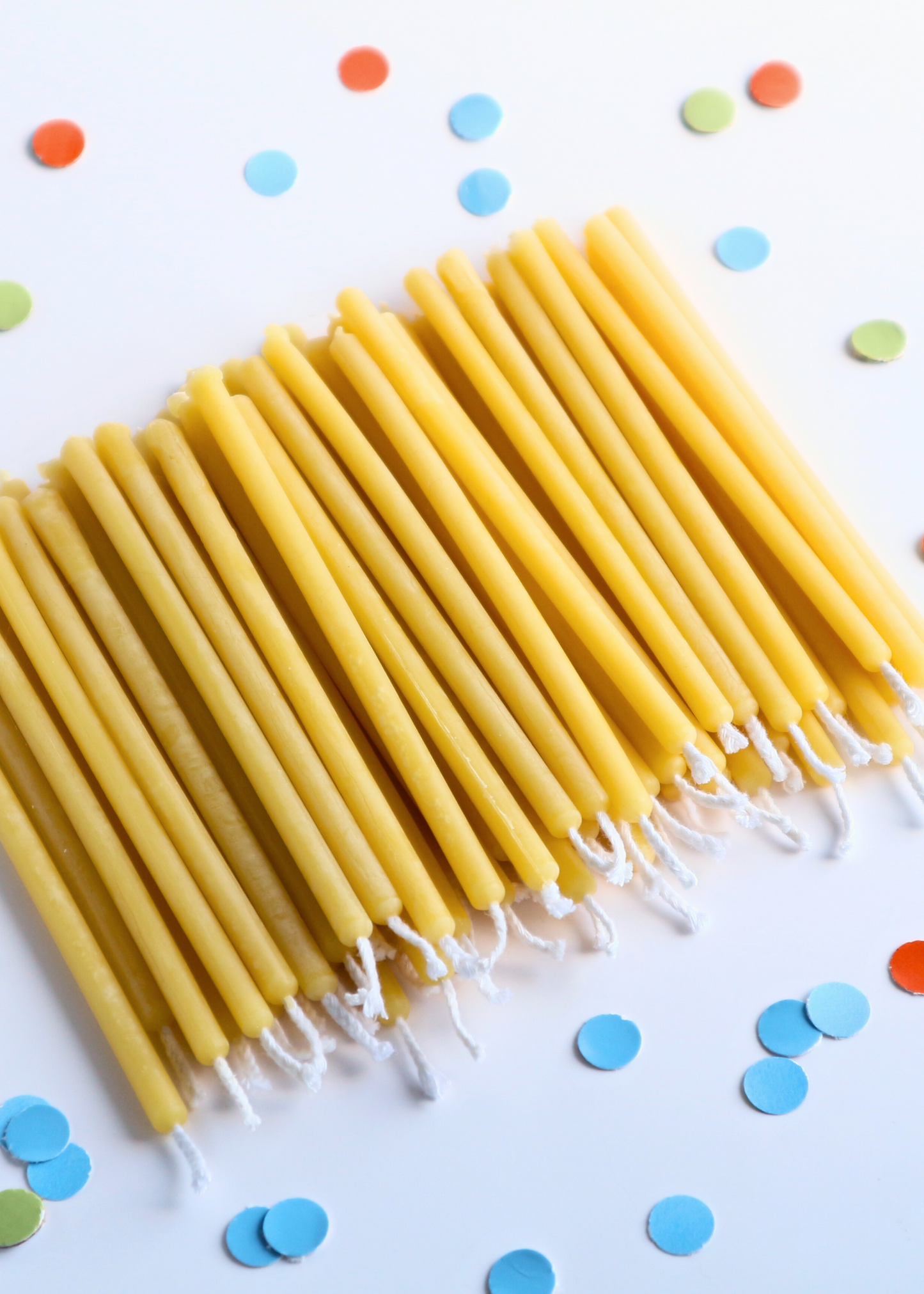 A close-up, overhead view of a hand holding a bundle of five 100% pure beeswax taper birthday candles with a white background and colorful birthday sprinkles.