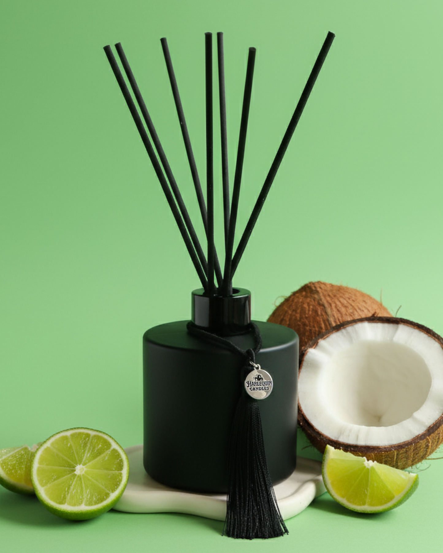 An Australian made Harlequin Candles black matte reed diffuser with a tropical coconut and lime scent called Coco Lime Sublime. The bottle is on a green background with sliced limes and cracked coconut.