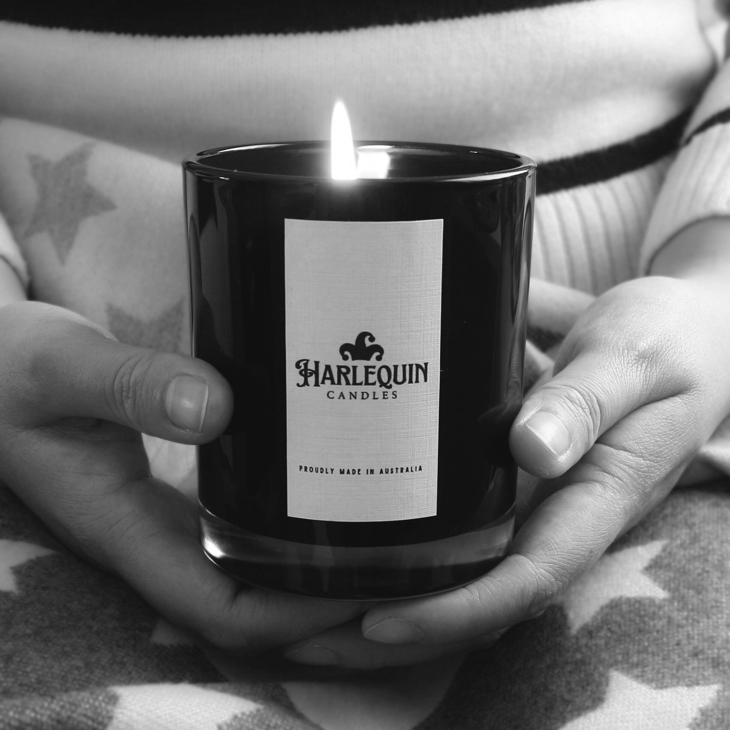 Monochrome image of hands gently cradling a lit Harlequin Candles soy candle in a matte black glass jar. The white label prominently displays the brand logo and 'PROUDLY MADE IN AUSTRALIA' text, set against a soft, patterned blanket, conveying warmth and quality craftsmanship.