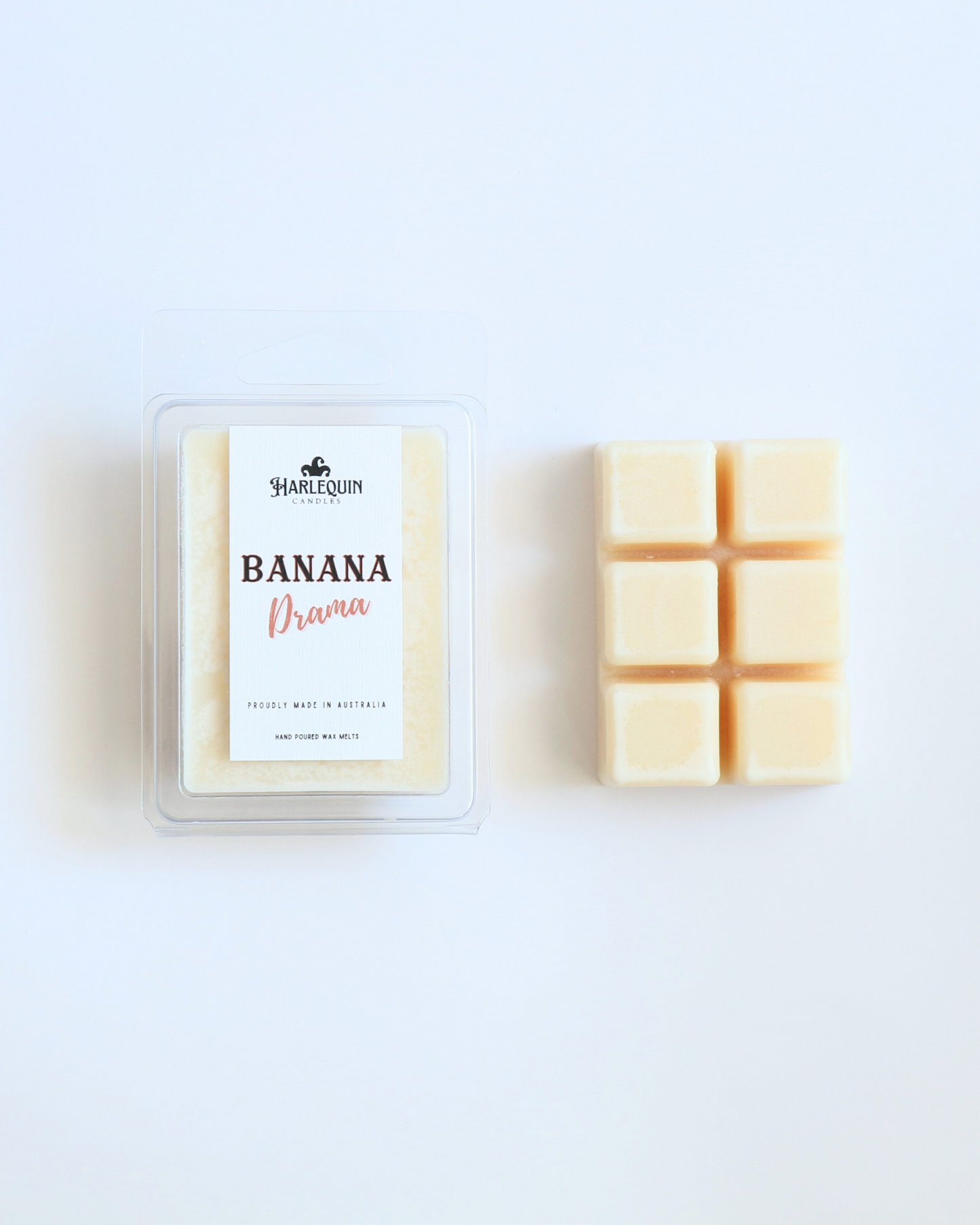 Harlequin Candles Banana Drama soy wax melts, displayed in clear plastic clamshell packaging with the brand label. The label reads 'BANANA Drama' and 'PROUDLY MADE IN AUSTRALIA HAND POURED WAX MELTS'. Several individual wax melt cubes are scattered on a light wooden cutting board, showcasing the fruity home fragrance product and its local origin.