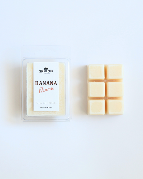 Harlequin Candles Banana Drama soy wax melts, displayed in clear plastic clamshell packaging with the brand label. The label reads 'BANANA Drama' and 'PROUDLY MADE IN AUSTRALIA HAND POURED WAX MELTS'. Several individual wax melt cubes are scattered on a light wooden cutting board, showcasing the fruity home fragrance product and its local origin.