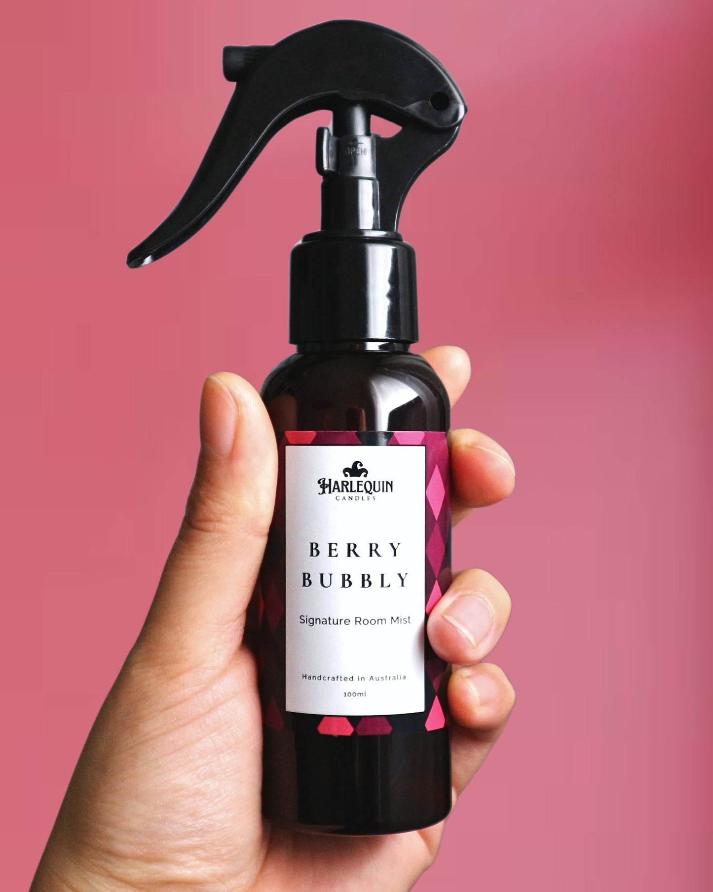 A Harlequin Candles Berry Bubbly Signature Room Mist in an amber spray bottle with a black trigger cap, placed against a two-toned background of light pink and white. The label clearly shows 'Harlequin Candles', 'Berry Bubbly', 'Signature Room Mist', and 'Handcrafted in Australia', highlighting this vibrant Australian-made home spray.