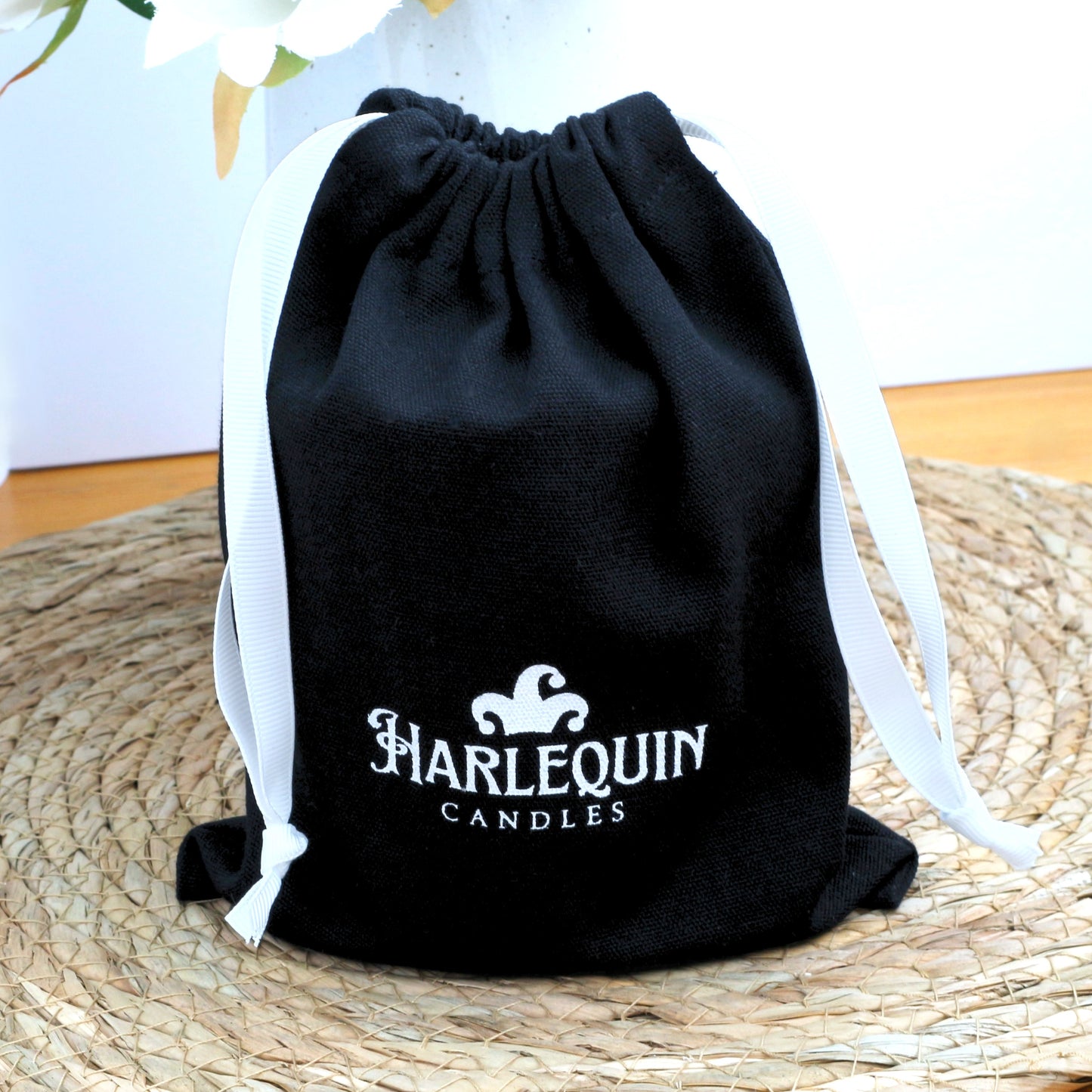 A black Harlequin Candles canvas drawstring gift bag with white ties, featuring the white Harlequin Candles logo. The reusable bag sits on a woven mat, with white flowers in a vase in the background, showcasing an elegant gifting option.
