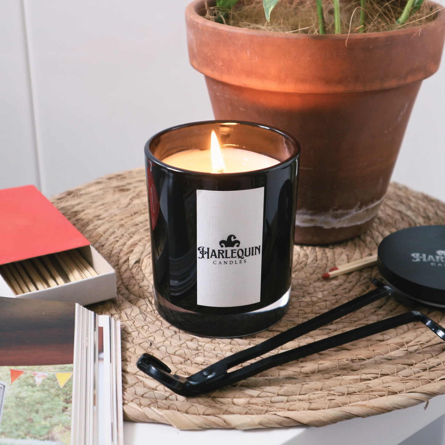 A lit Harlequin Candles soy candle in a matte black glass jar, resting on a rustic woven mat. Beside it are a red box of matches, a black candle wick trimmer, and a black lid. A terracotta potted plant is in the background, creating a warm and inviting Australian home ambiance.