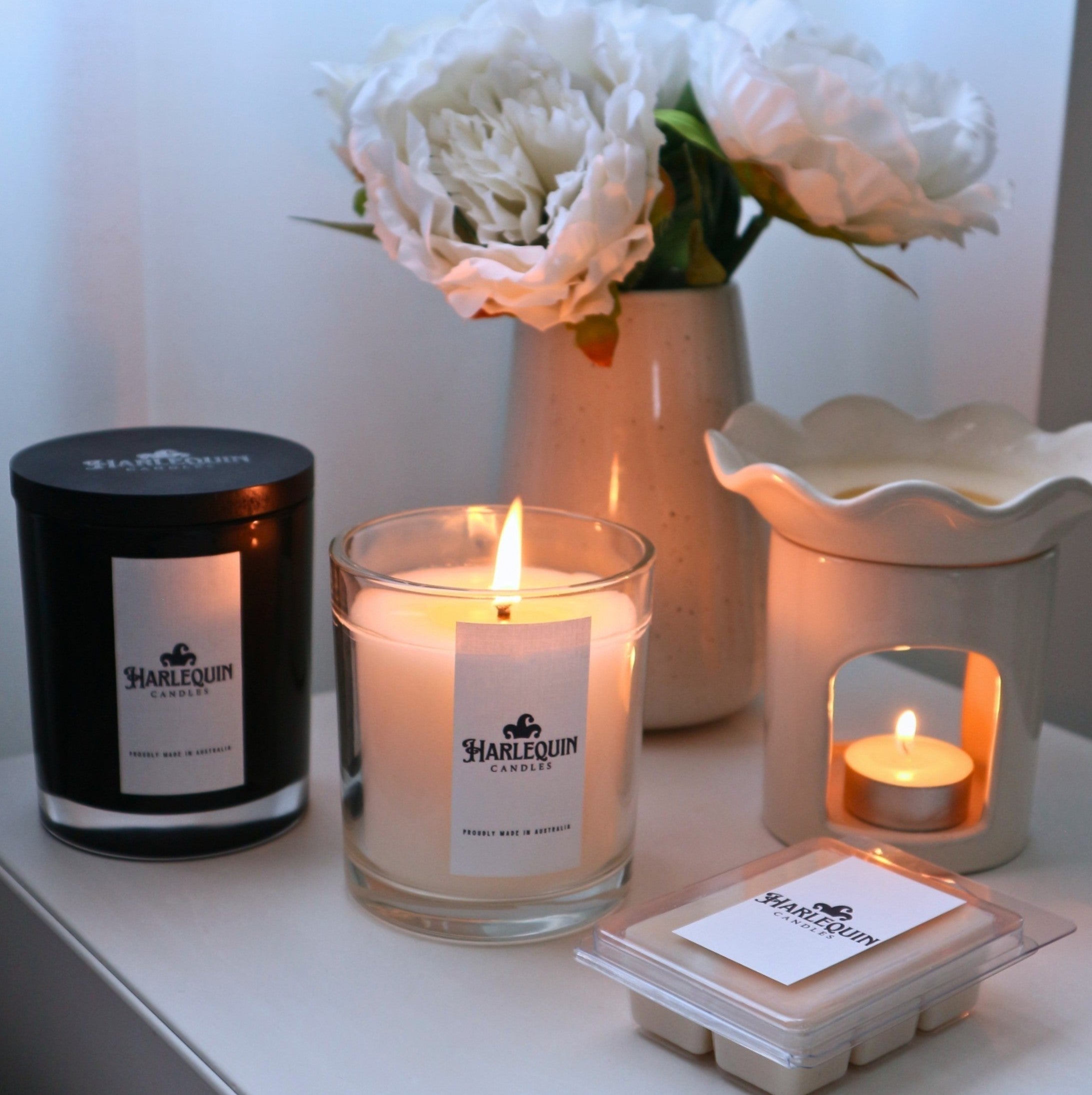 A curated display of Harlequin Candles home fragrance products, featuring a black jar soy candle, a lit soy candle in a clear glass jar (showing 'Proudly Made in Australia' on the label), a white ceramic wax warmer with a lit tealight, and a clamshell of soy wax melts. A vase with white faux peonies sits in the background, creating a cozy and inviting atmosphere.