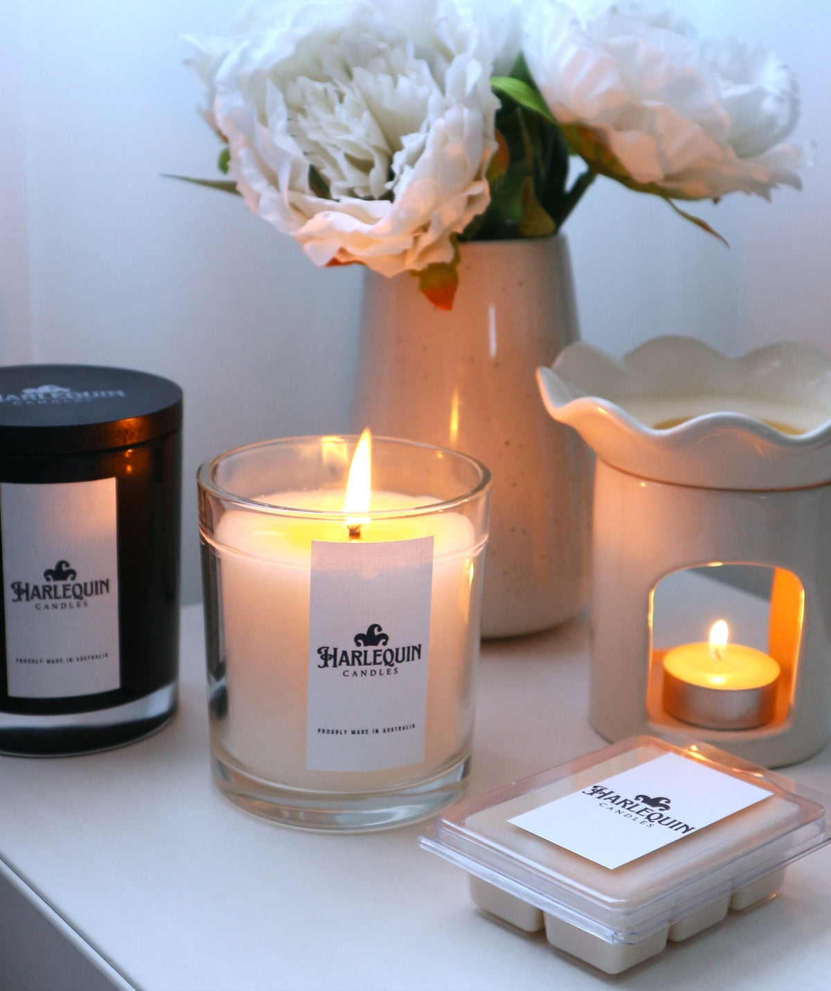 A curated display of Harlequin Candles home fragrance products, featuring a black jar soy candle, a lit soy candle in a clear glass jar (showing 'Proudly Made in Australia' on the label), a white ceramic wax warmer with a lit tealight, and a clamshell of soy wax melts. A vase with white faux peonies sits in the background, creating a cozy and inviting atmosphere.