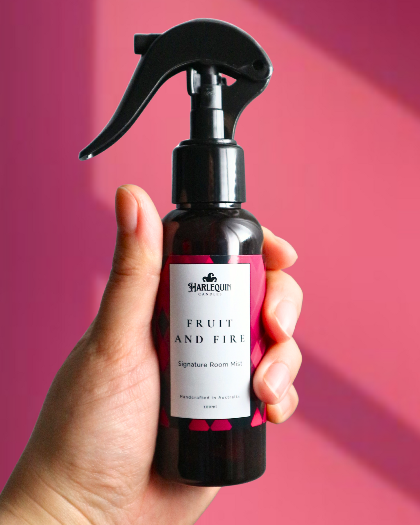 A Harlequin Candles Dragonfruit, Chilli, Chipotle inspired Signature Room Mist in an amber spray bottle with a black trigger cap, placed against a two-toned background of pink and red. The label clearly shows 'Harlequin Candles', 'Fruit and Fire', 'Signature Room Mist', and 'Handcrafted in Australia', highlighting this enchanting Australian-made home spray.