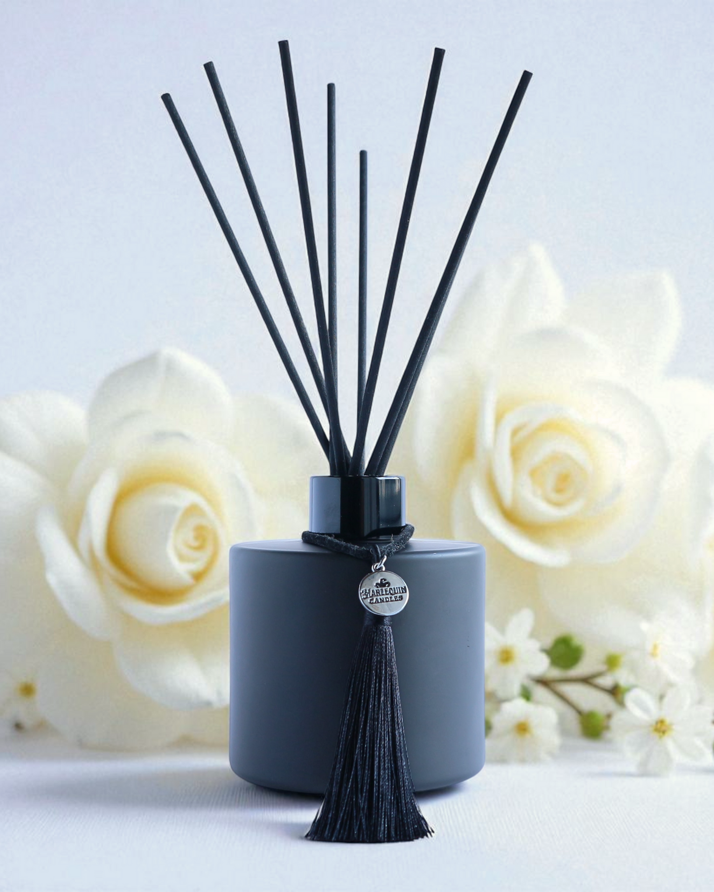 Harlequin Candles Gardenia Bloom reed diffuser in a matte black bottle with black reeds and a decorative black tassel with a silver logo charm. A large, pristine white gardenia flower is next to the diffuser, set against a soft purple background, evoking a lush and elegant home fragrance.