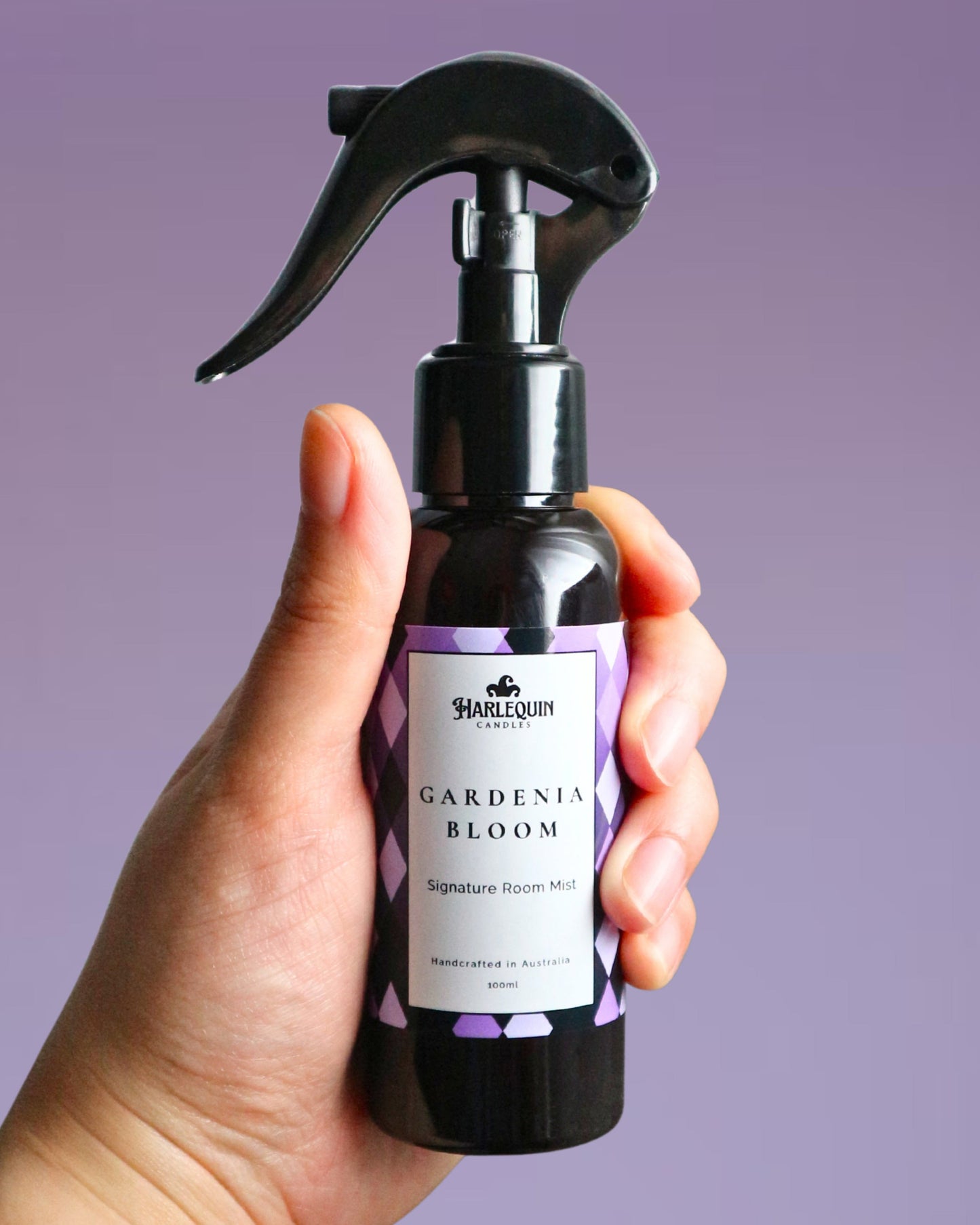 A Harlequin Candles Gardenia Bloom Signature Room Mist in an amber spray bottle with a black trigger cap, placed against a two-toned background of light pink and white. The label clearly shows 'Harlequin Candles', 'Gardenia Bloom', 'Signature Room Mist', and 'Handcrafted in Australia', highlighting this lush floral Australian-made home spray.