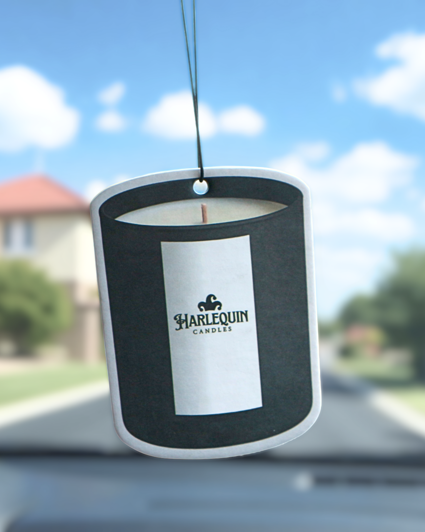 A Harlequin Candles candle-shaped hanging car freshener with a white label prominently featuring the brand logo, suspended from a car's rearview mirror. The interior of the car is visible, with blurred trees and buildings through the windshield, showcasing a stylish Australian car air freshener.