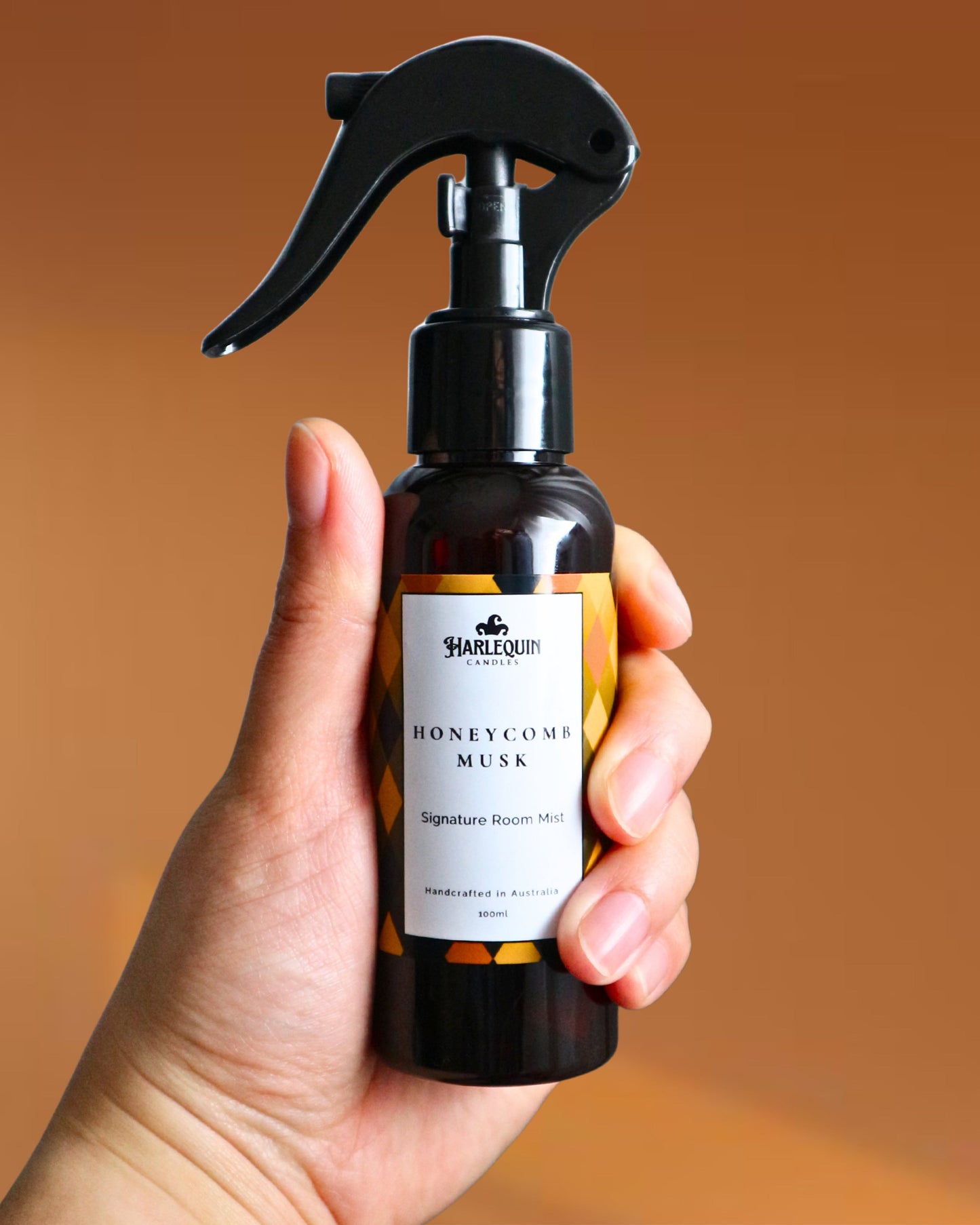 A Harlequin Candles Honeycomb Musk Signature Room Mist in an amber spray bottle with a black trigger cap, placed against a two-toned background of light yellow and white. The label clearly shows 'Harlequin Candles', 'Honeycomb Musk', 'Signature Room Mist', and 'Handcrafted in Australia', highlighting this warm and comforting Australian-made home spray.