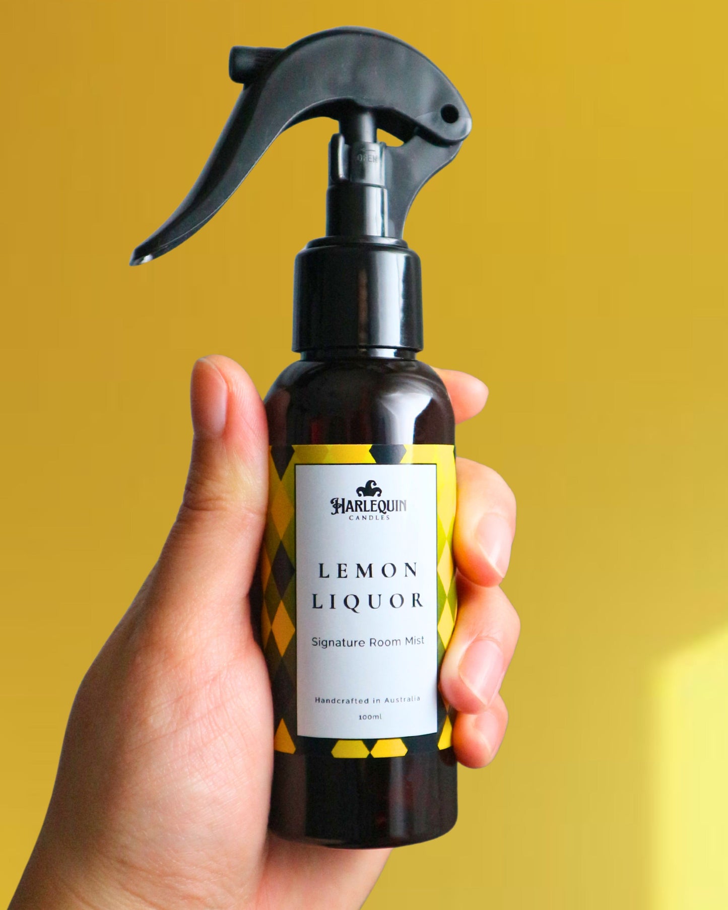 A Harlequin Candles Lemon Liquor (Limoncello scented) Signature Room Mist in an amber spray bottle with a black trigger cap, placed against a two-toned background of light yellow and white. The label clearly shows 'Harlequin Candles', 'Lemon Liquor', 'Signature Room Mist', and 'Handcrafted in Australia', highlighting this zesty Australian-made home spray.