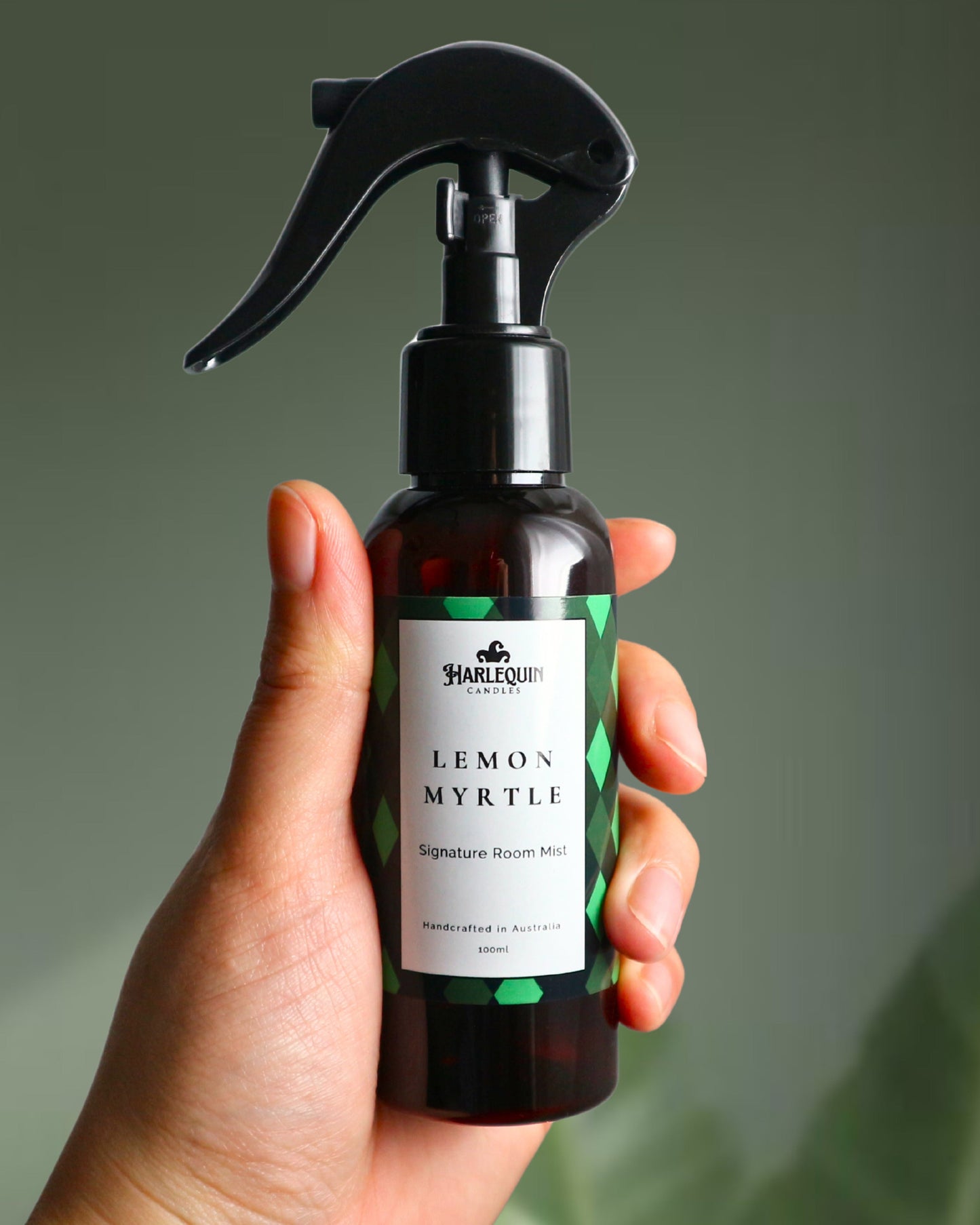 A Harlequin Candles Lemon Myrtle Signature Room Mist in an amber spray bottle with a black trigger cap, placed against a two-toned background of light green and white. The label clearly shows 'Harlequin Candles', 'Lemon Myrtle', 'Signature Room Mist', and 'Handcrafted in Australia', highlighting this refreshing Australian-made home spray.