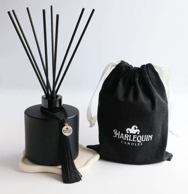 A sleek matte black Harlequin Candles reed diffuser bottle with black reeds, a black tassel charm, and a black drawstring gift bag featuring the white Harlequin Candles logo. The items are artfully arranged on a subtle white coaster against a clean white background, showcasing a luxury home fragrance product.