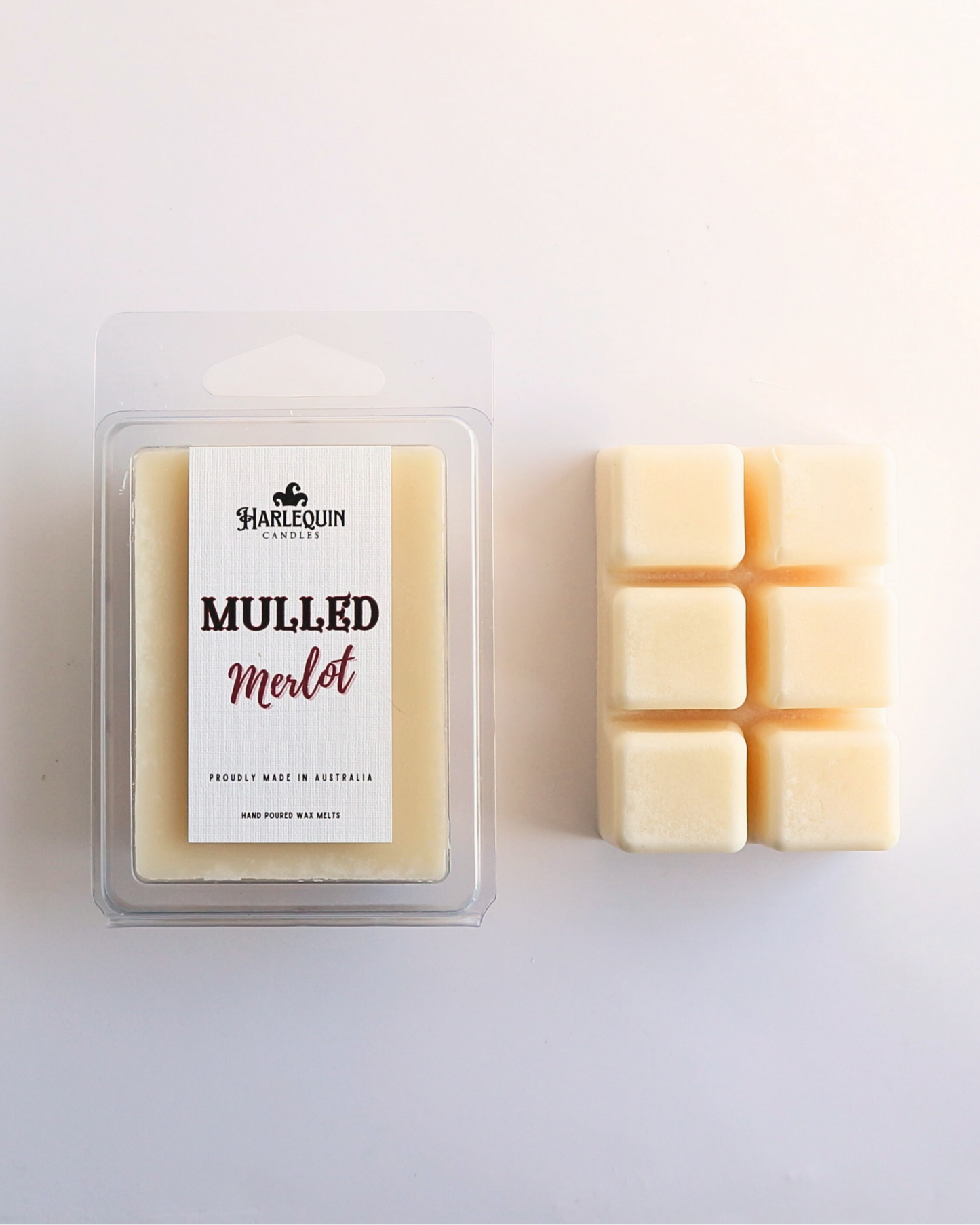 Harlequin Candles Mulled Merlot soy wax melts, displayed in clear plastic clamshell packaging with the brand label. The label reads 'MULLED Merlot' and 'PROUDLY MADE IN AUSTRALIA HAND POURED WAX MELTS'. Several individual wax melt cubes are scattered on a light wooden cutting board, showcasing the cozy winter home fragrance product and its local origin.