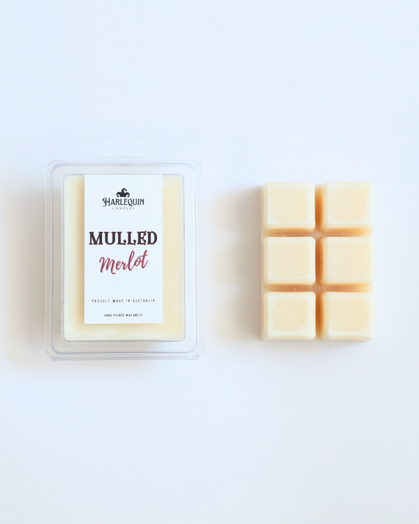Harlequin Candles Mulled Merlot soy wax melts, displayed in clear plastic clamshell packaging with the brand label. The label reads 'MULLED Merlot' and 'PROUDLY MADE IN AUSTRALIA HAND POURED WAX MELTS'. Several individual wax melt cubes are scattered on a light wooden cutting board, showcasing the cozy winter home fragrance product and its local origin.