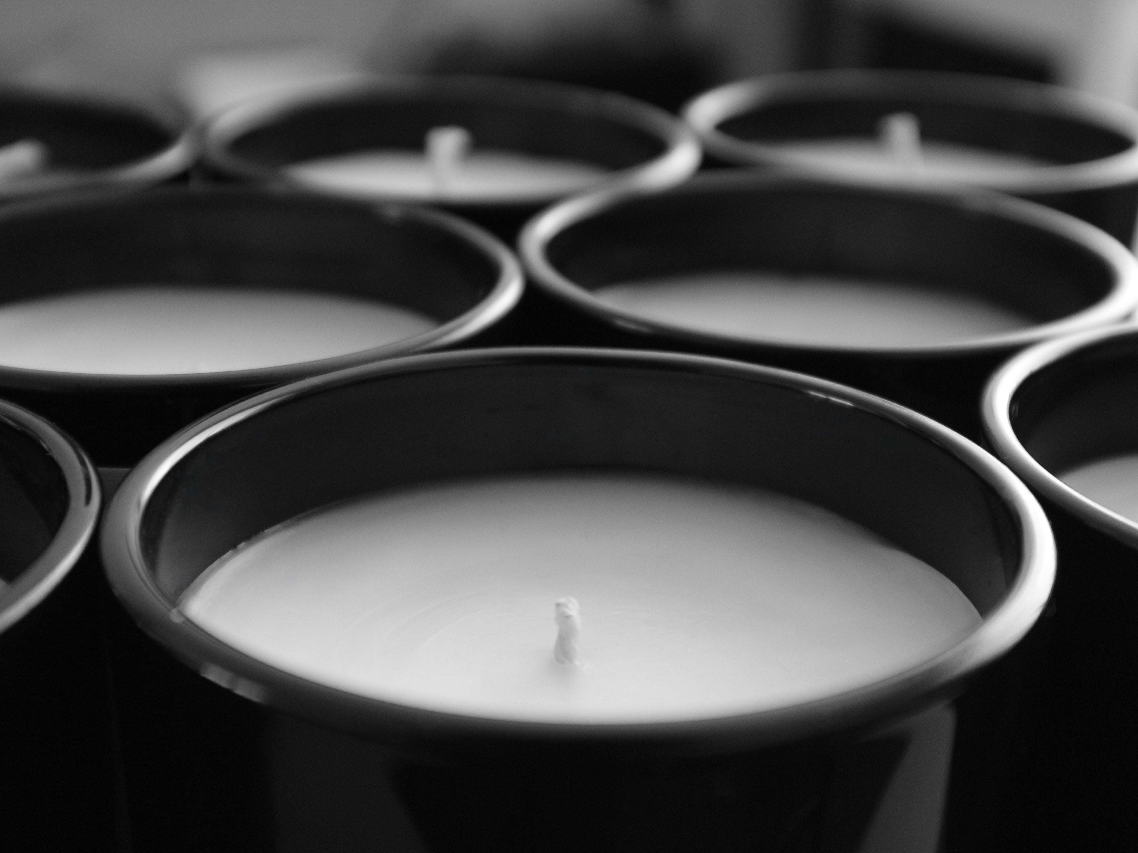 Close-up, monochrome view of multiple unlit Harlequin Candles in sleek black glass jars, showing their white wax and wicks in a repeating pattern. The image conveys the minimalist and luxurious aesthetic of the candle collection.