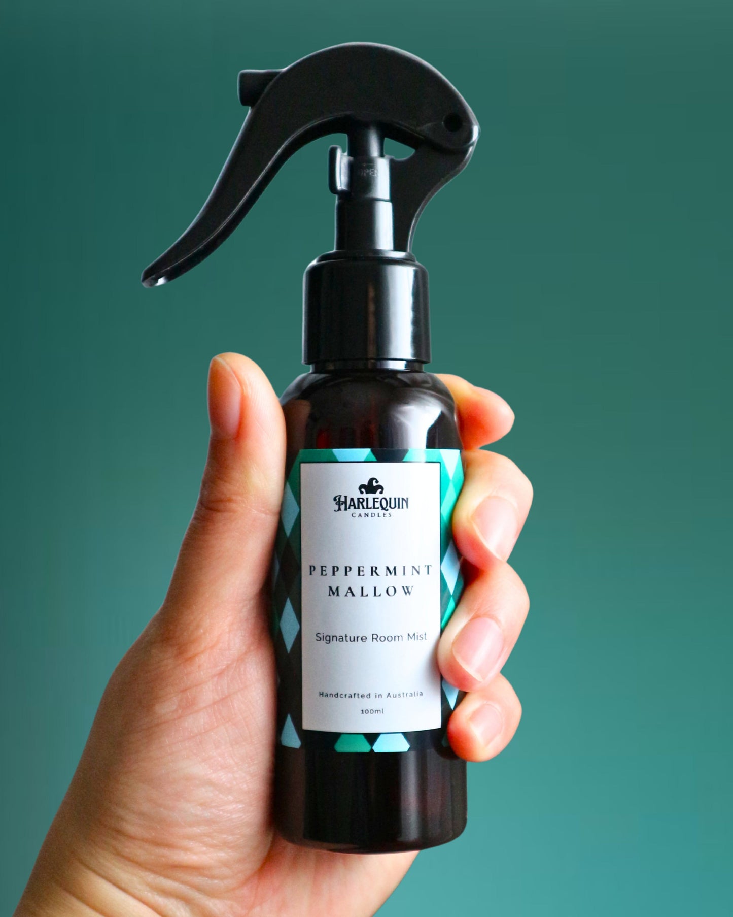 A Harlequin Candles Peppermint Mallow Signature Room Mist in an amber spray bottle with a black trigger cap, placed against a two-toned background of light blue and white. The label clearly shows 'Harlequin Candles', 'Peppermint Mallow', 'Signature Room Mist', and 'Handcrafted in Australia', highlighting this sweet mint Australian-made home spray.