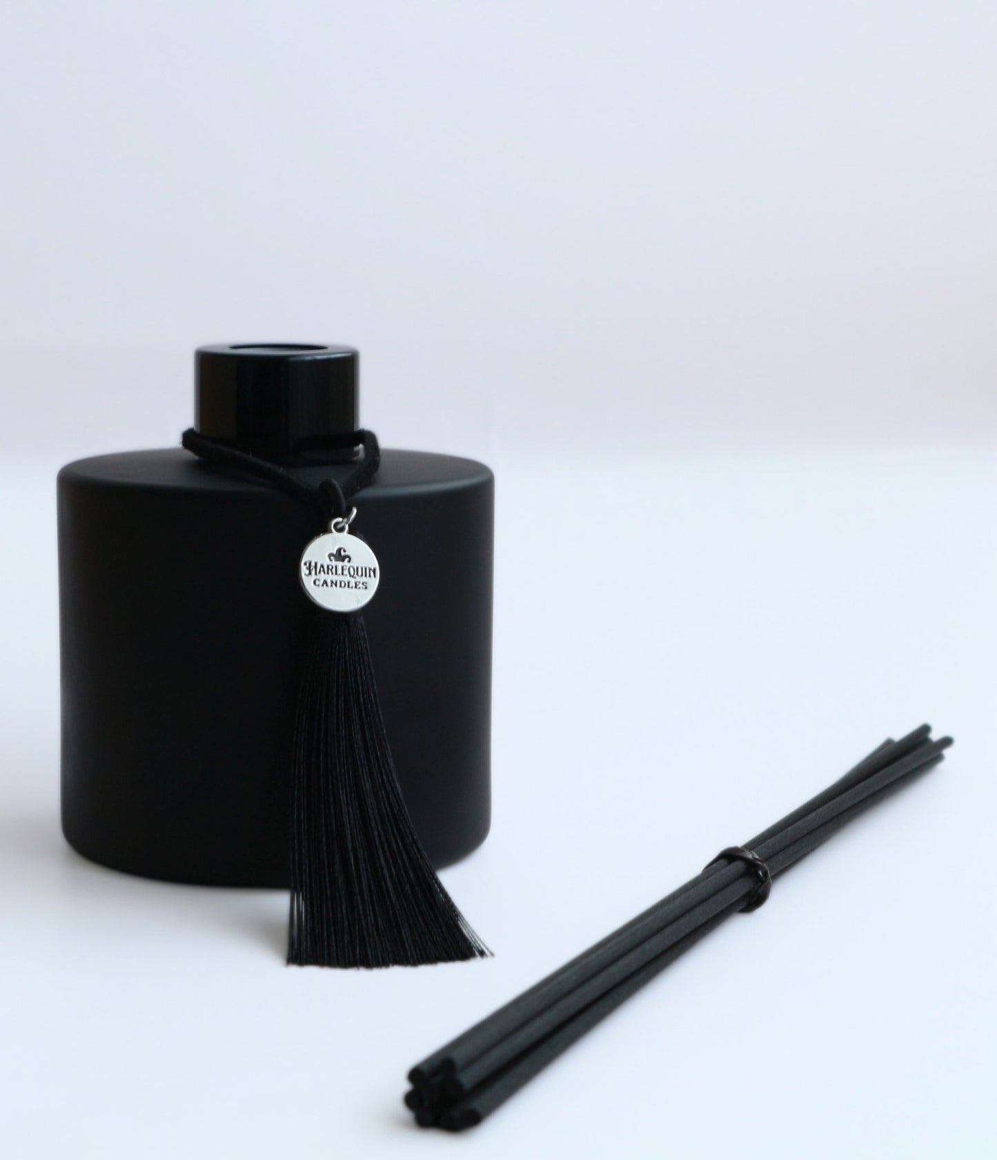 A Harlequin Candles premium matte black reed diffuser bottle with a black tassel and silver logo charm, displayed next to a bundle of black diffuser reeds, all on a clean white background. Showcases a stylish home fragrance diffuser set.
