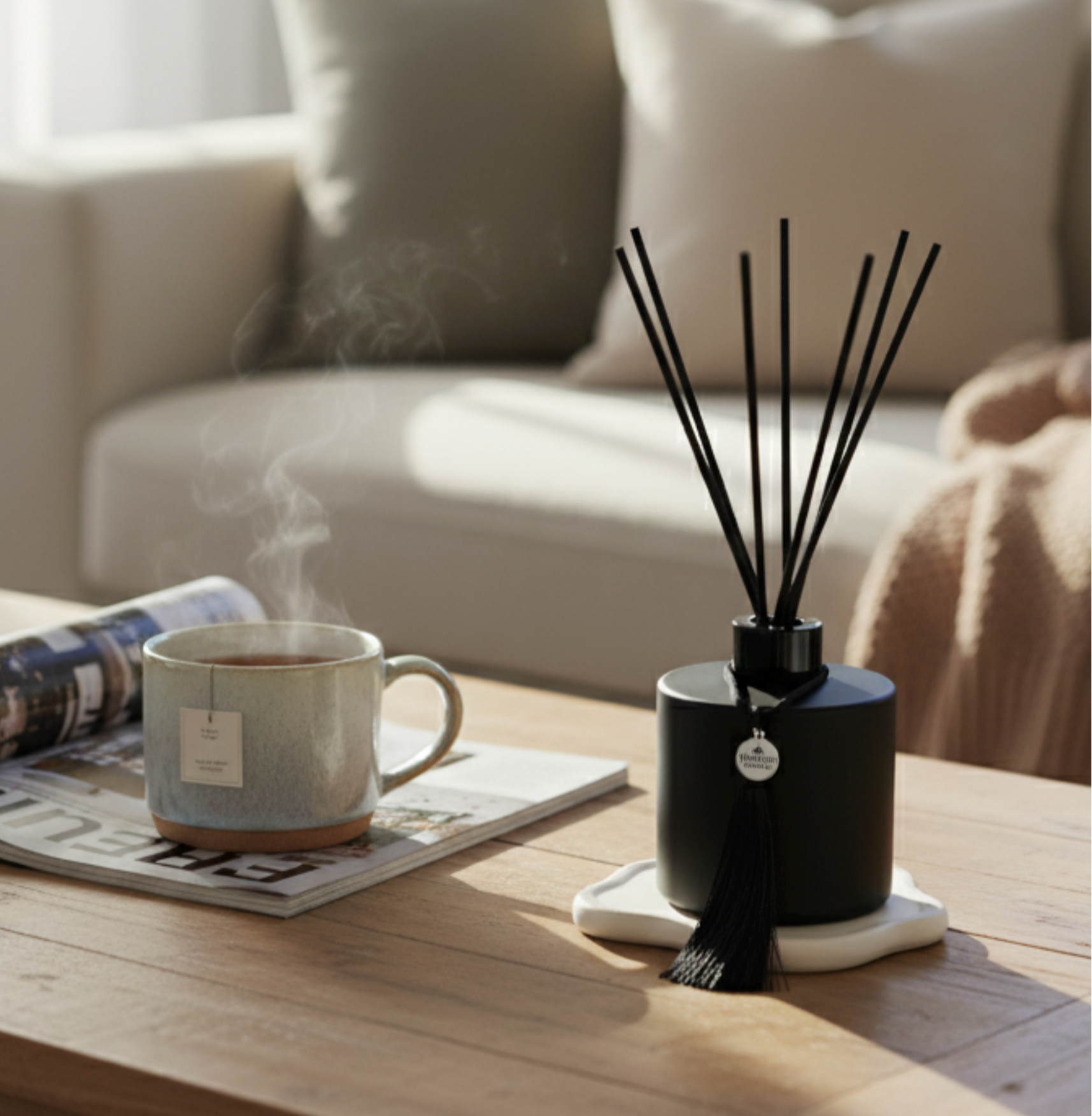 A Harlequin Candles matte black reed diffuser with black reeds and a tassel charm sits on a white coaster on a wooden coffee table in a warm, inviting living room. Next to it, a steaming mug of tea rests on an open magazine. A light-colored sofa with throw pillows and a blanket is visible in the softly lit background, creating an atmosphere of luxury and comfort.