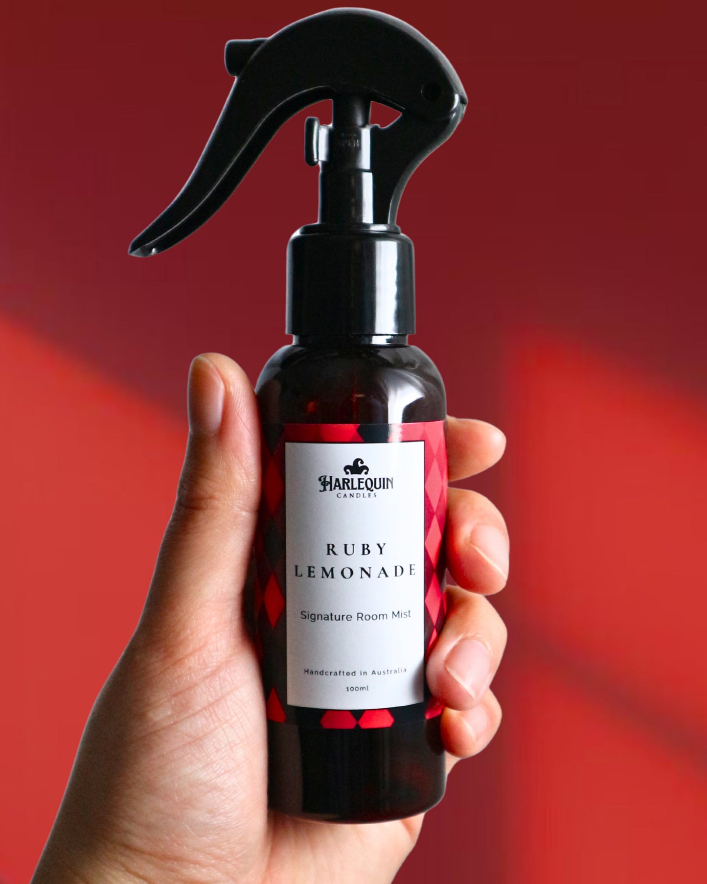 A Harlequin Candles Ruby Lemonade Signature Room Mist in an amber spray bottle with a black trigger cap, placed against a two-toned background of light pink and white. The label clearly shows 'Harlequin Candles', 'Ruby Lemonade', 'Signature Room Mist', and 'Handcrafted in Australia', highlighting this vibrant Australian-made home spray.