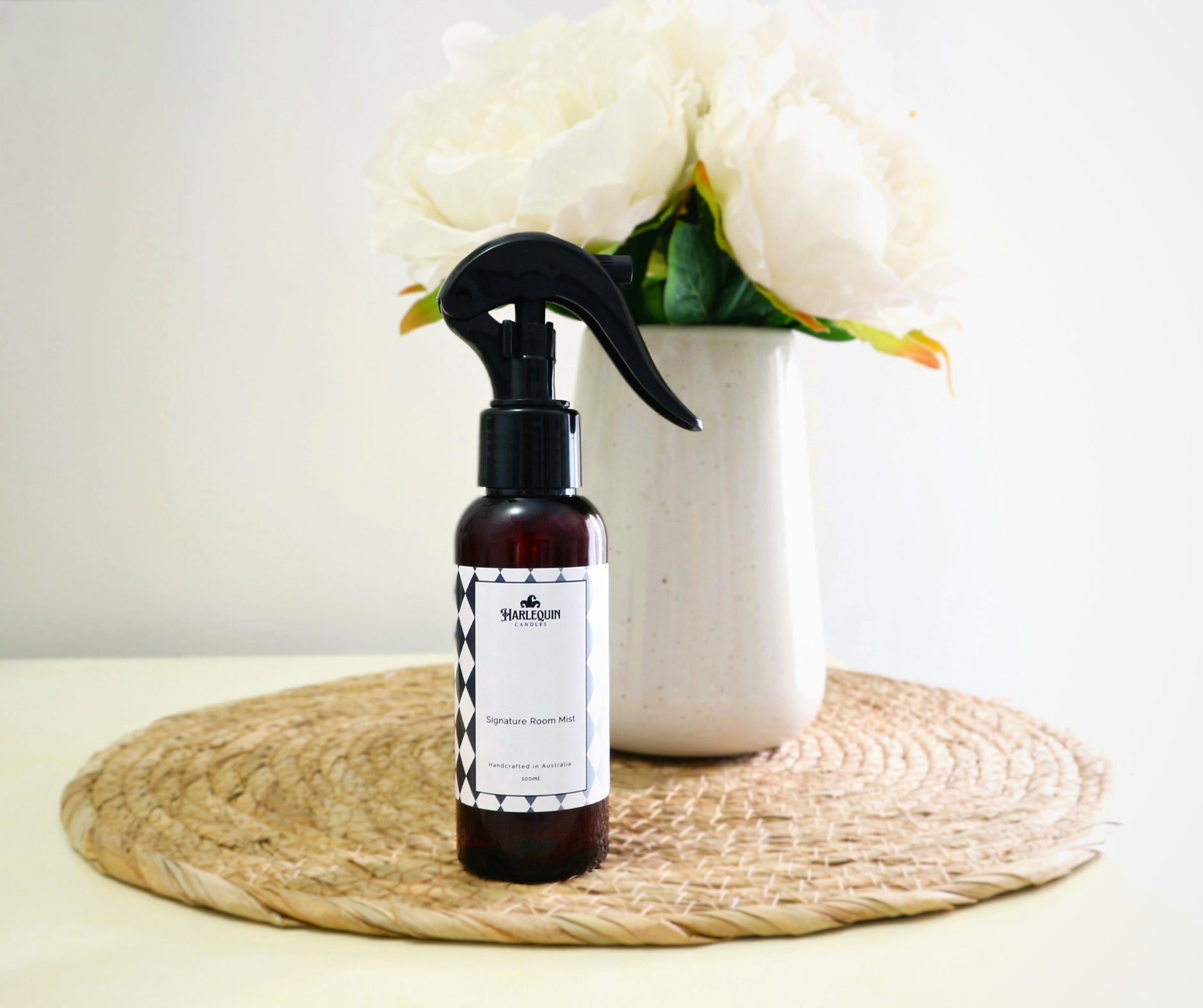 Harlequin Candles Signature Room Mist in an amber spray bottle with a black trigger cap, placed on a light woven mat. In the background, a white vase holds white faux peonies, conveying a fresh and elegant home fragrance atmosphere.