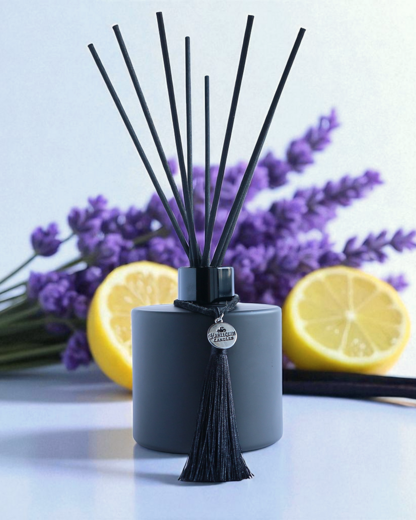 Harlequin Candles Vanilla Lavender Love Affair reed diffuser in a matte black bottle with black reeds and a decorative black tassel with a silver logo charm. A sprig of purple lavender lies beside the diffuser on a vibrant purple background, evoking a soothing and elegant home fragrance.