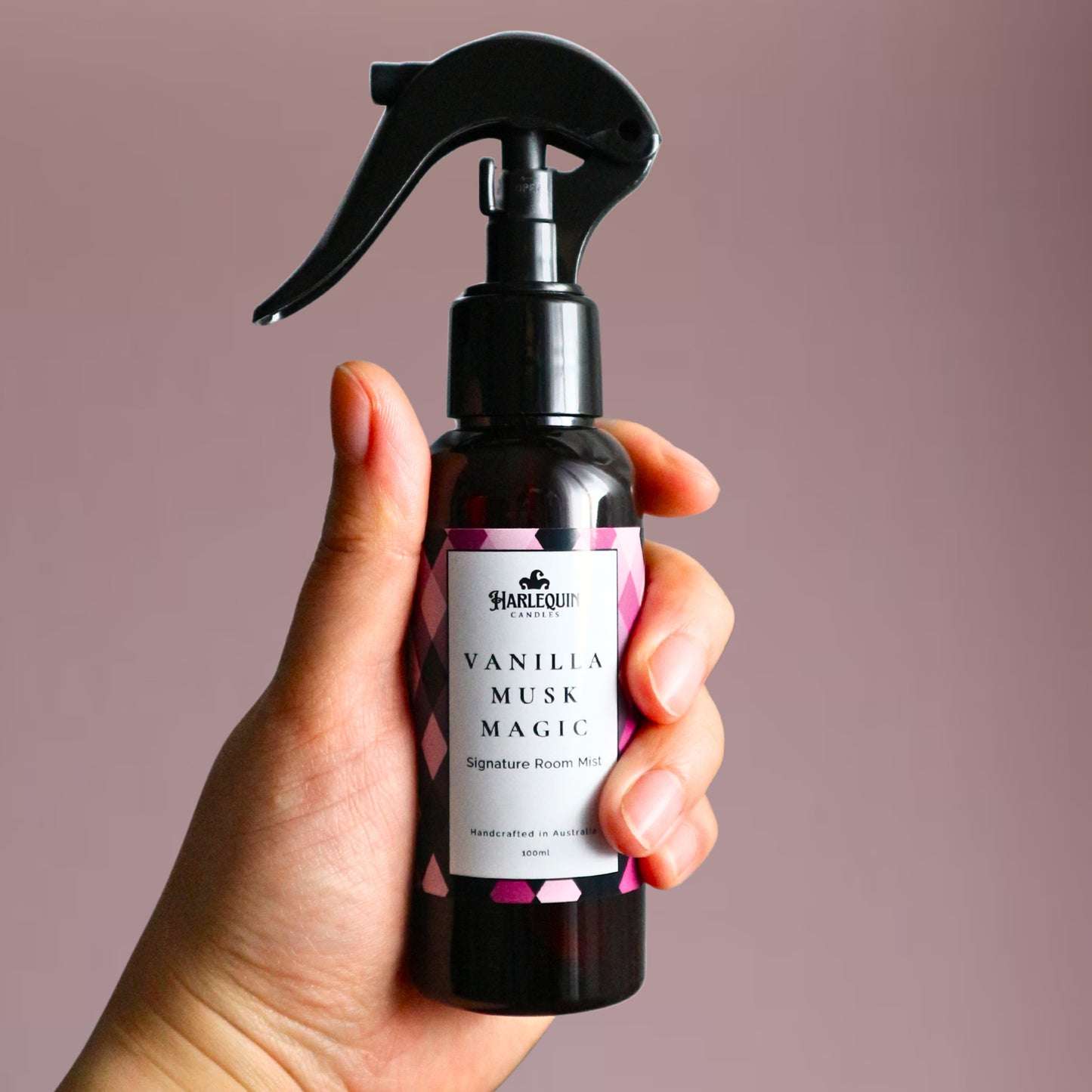 A Harlequin Candles Vanilla Musk Magic Signature Room Mist in an amber spray bottle with a black trigger cap, placed against a two-toned background of light pink and white. The label clearly shows 'Harlequin Candles', 'Vanilla Musk Magic', 'Signature Room Mist', and 'Handcrafted in Australia', highlighting this enchanting Australian-made home spray.