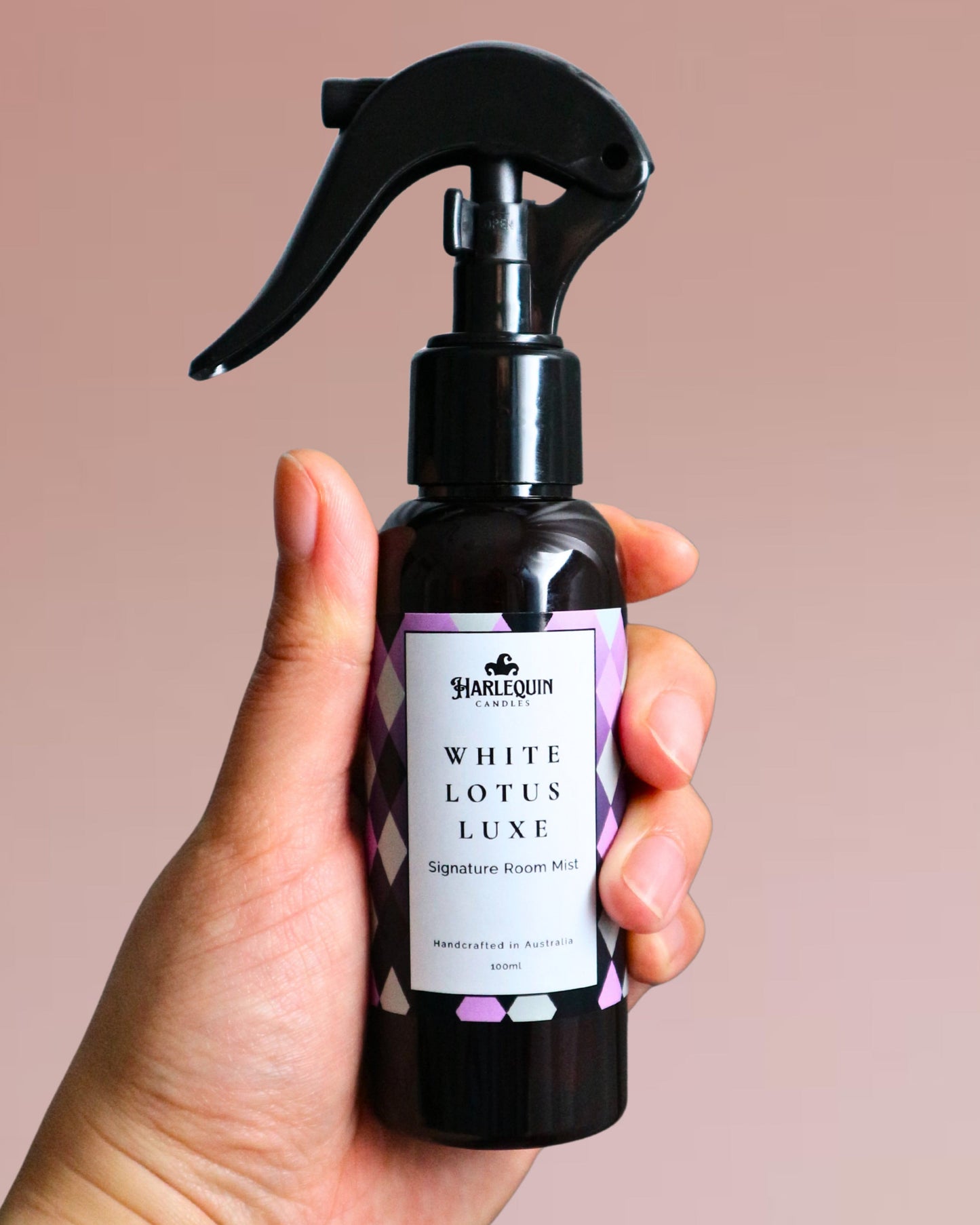A Harlequin Candles White Lotus Luxe Signature Room Mist in an amber spray bottle with a black trigger cap, placed against a two-toned background of light blue and white. The label clearly shows 'Harlequin Candles', 'White Lotus Luxe', 'Signature Room Mist', and 'Handcrafted in Australia', highlighting this elegant Australian-made home spray.