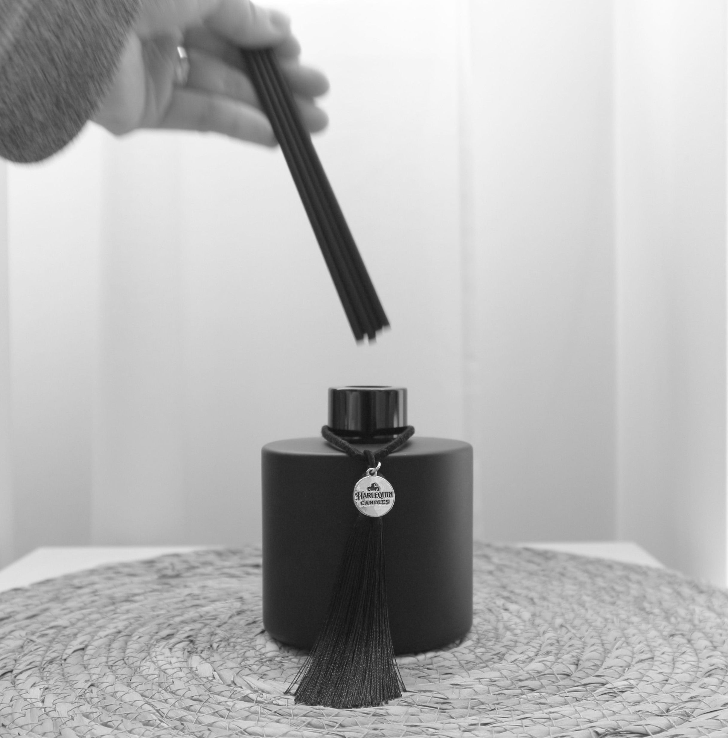 Close-up of a hand inserting black reeds into a matte black Harlequin Candles reed diffuser bottle, which features a decorative black tassel and silver logo charm, demonstrating the setup process for a new home fragrance diffuser.