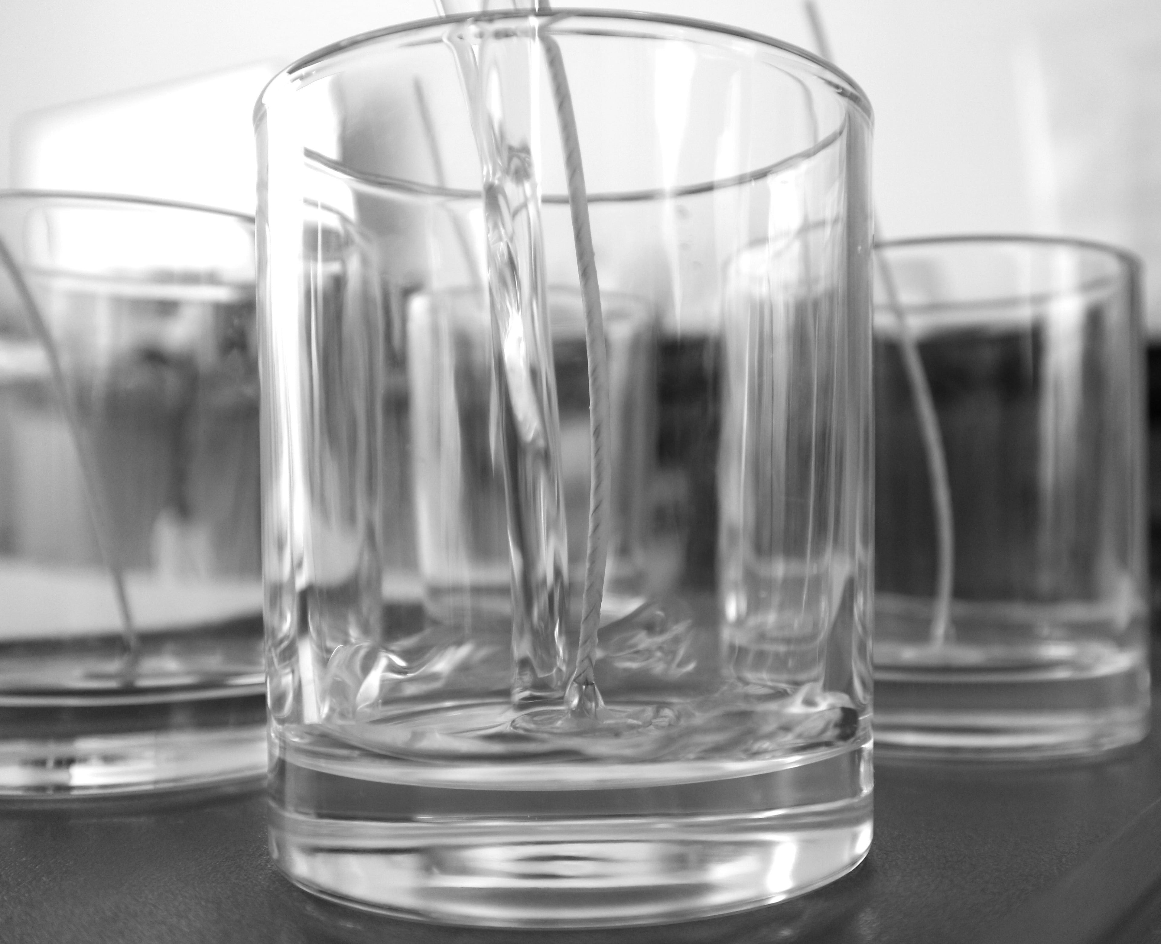 Monochrome close-up of clear liquid being precisely poured into a clean, empty glass tumbler, with other empty glasses blurred in the background. Illustrates the careful pouring and pure ingredients used in Harlequin Candles' products.