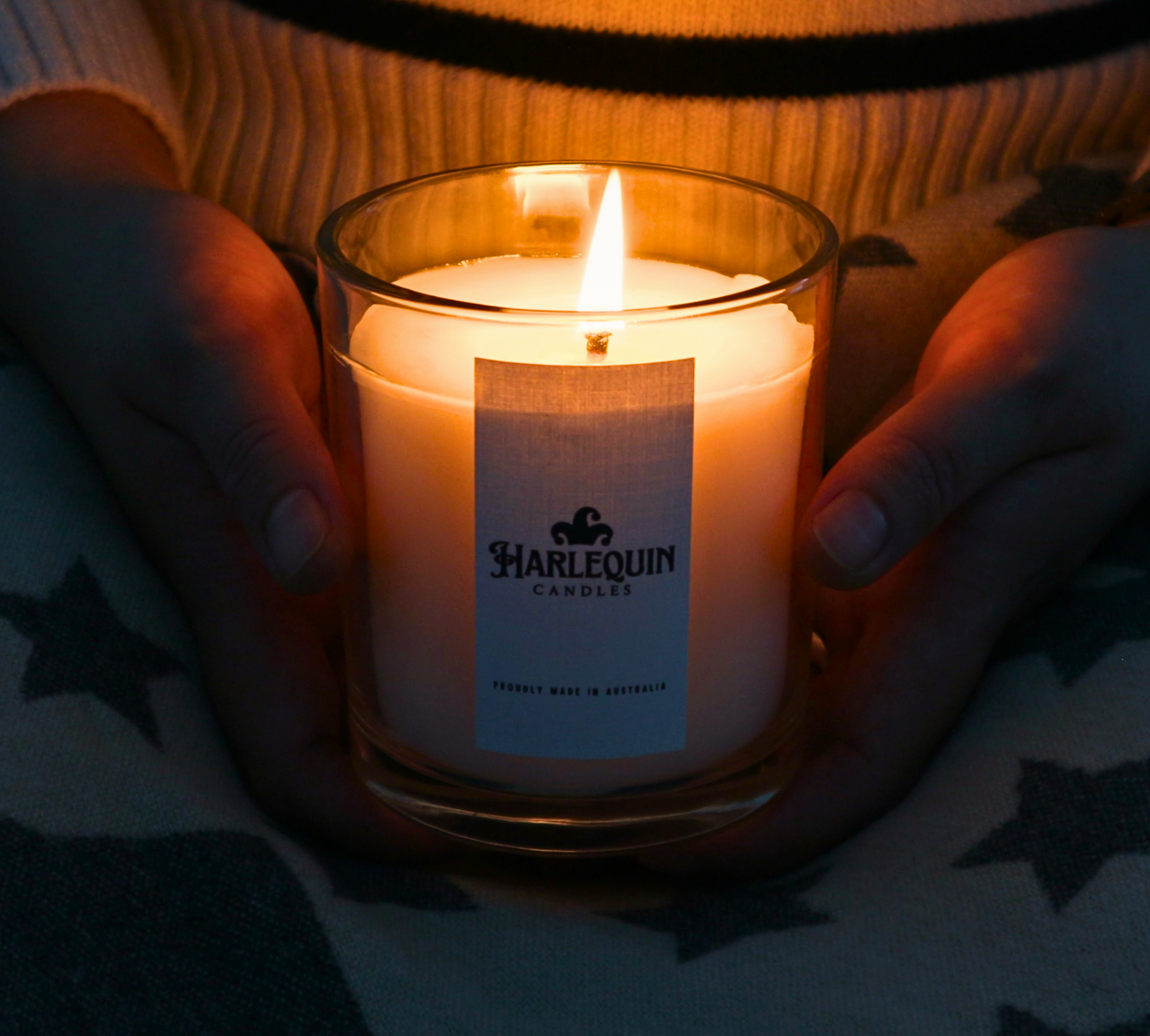 A person's hands, wearing a cozy striped sweater, gently cup a lit Harlequin Candles scented candle in a clear glass jar. The warm glow of the flame illuminates the candle and the surrounding hands. The white label on the candle jar features the Harlequin Candles logo and text, and a soft, blurred background shows a patterned blanket, enhancing the sense of comfort and warmth.