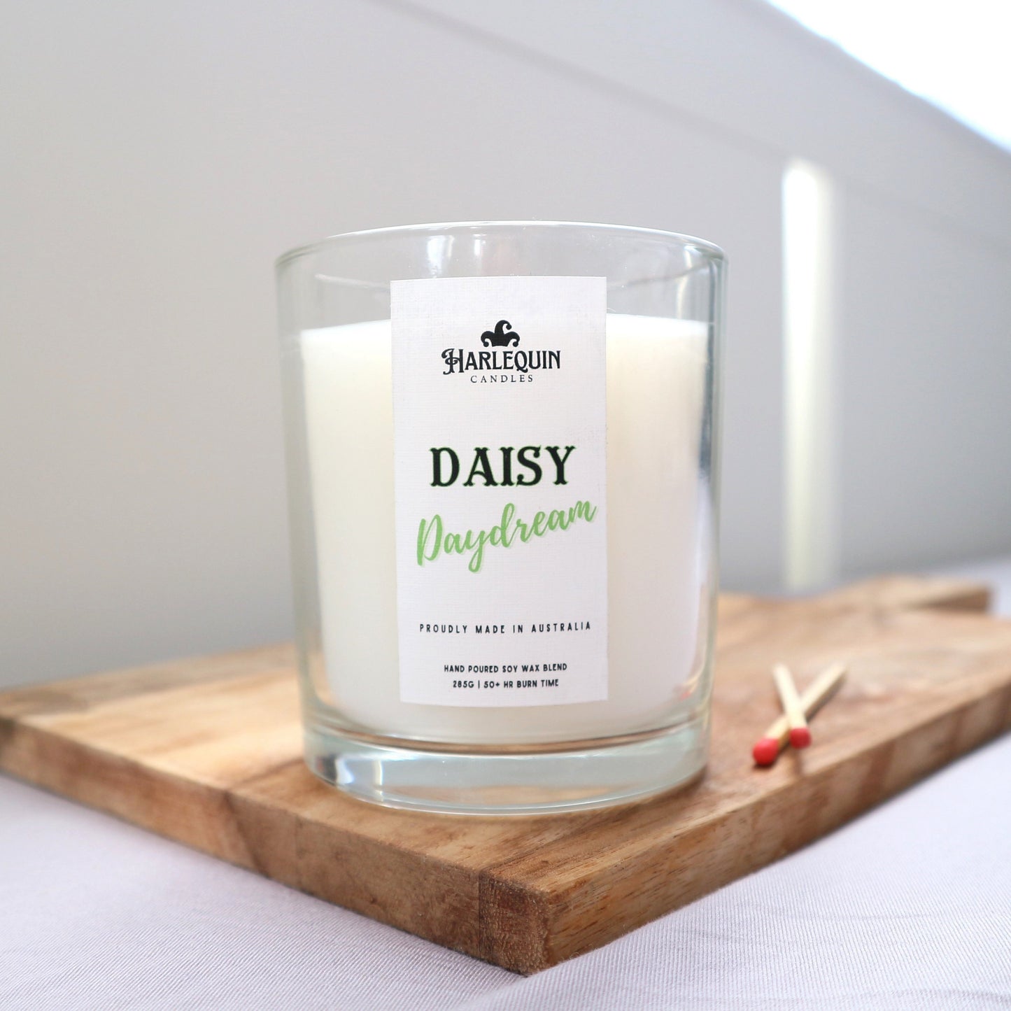 A Harlequin Candles "Daisy Daydream" hand-poured soy blend candle in a clear glass jar, displayed on a wooden board with matches. Proudly Australian made.