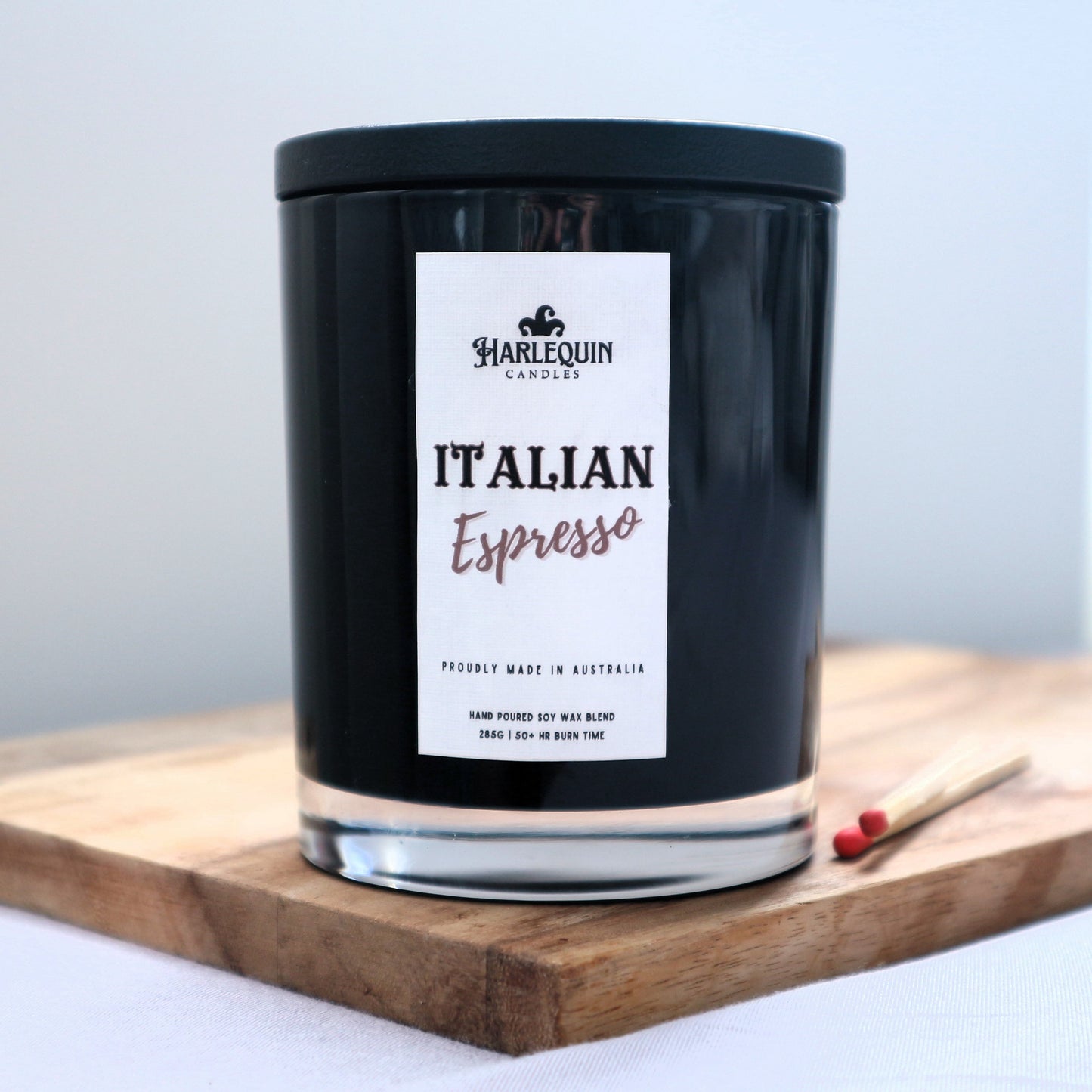 A Harlequin Candles "Italian Espresso" hand-poured soy blend candle in a sleek black glass jar with lid, displayed on a wooden board with matches. Proudly Australian made.