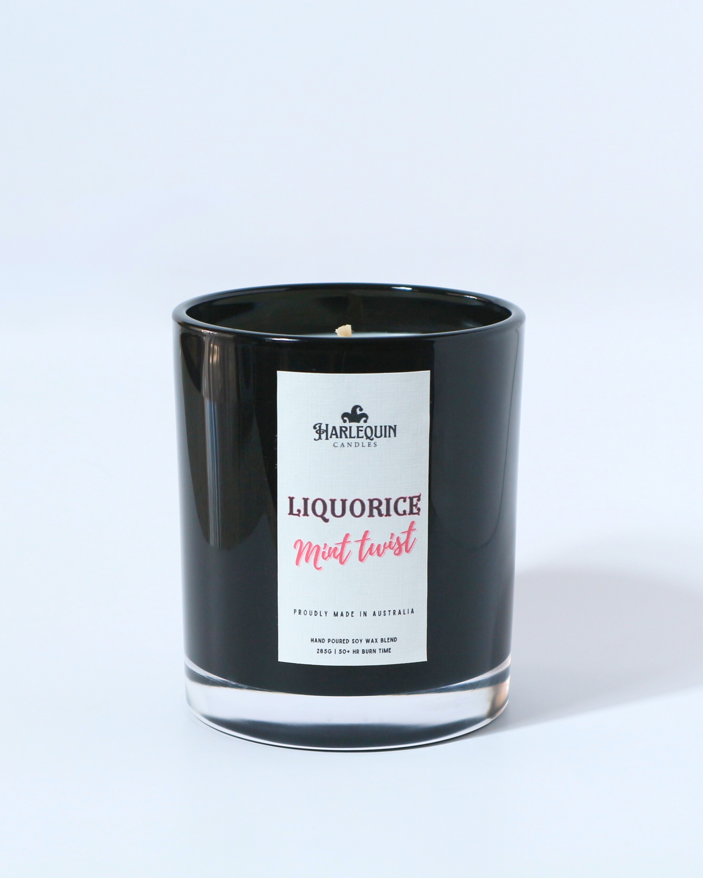 A Harlequin Candles "Liquorice Mint Twist" hand-poured soy wax candle in a black glass jar, styled beside a clear glass vase with vibrant pink roses. Premium Australian made scented candle.