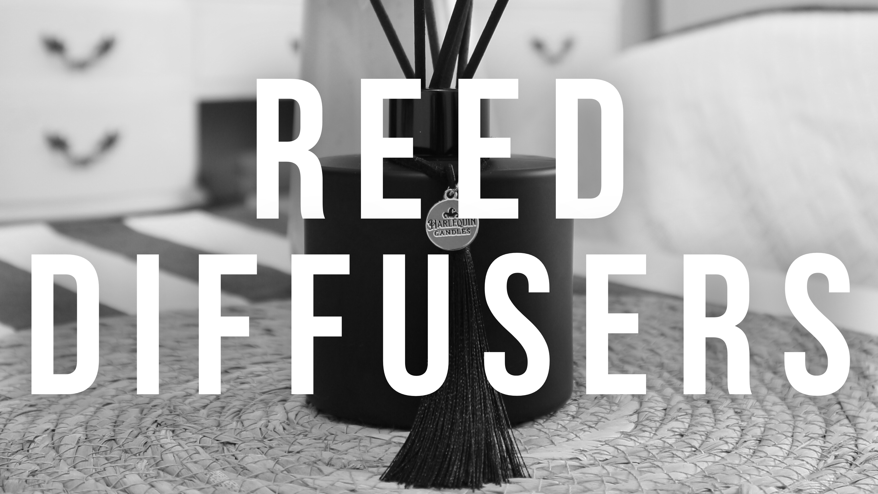 Monochrome banner with 'REED DIFFUSERS' text prominently displayed. Features a Harlequin Candles matte black reed diffuser with black reeds and tassel in the foreground, with a blurred vase of white flowers in the background, promoting premium home fragrance.