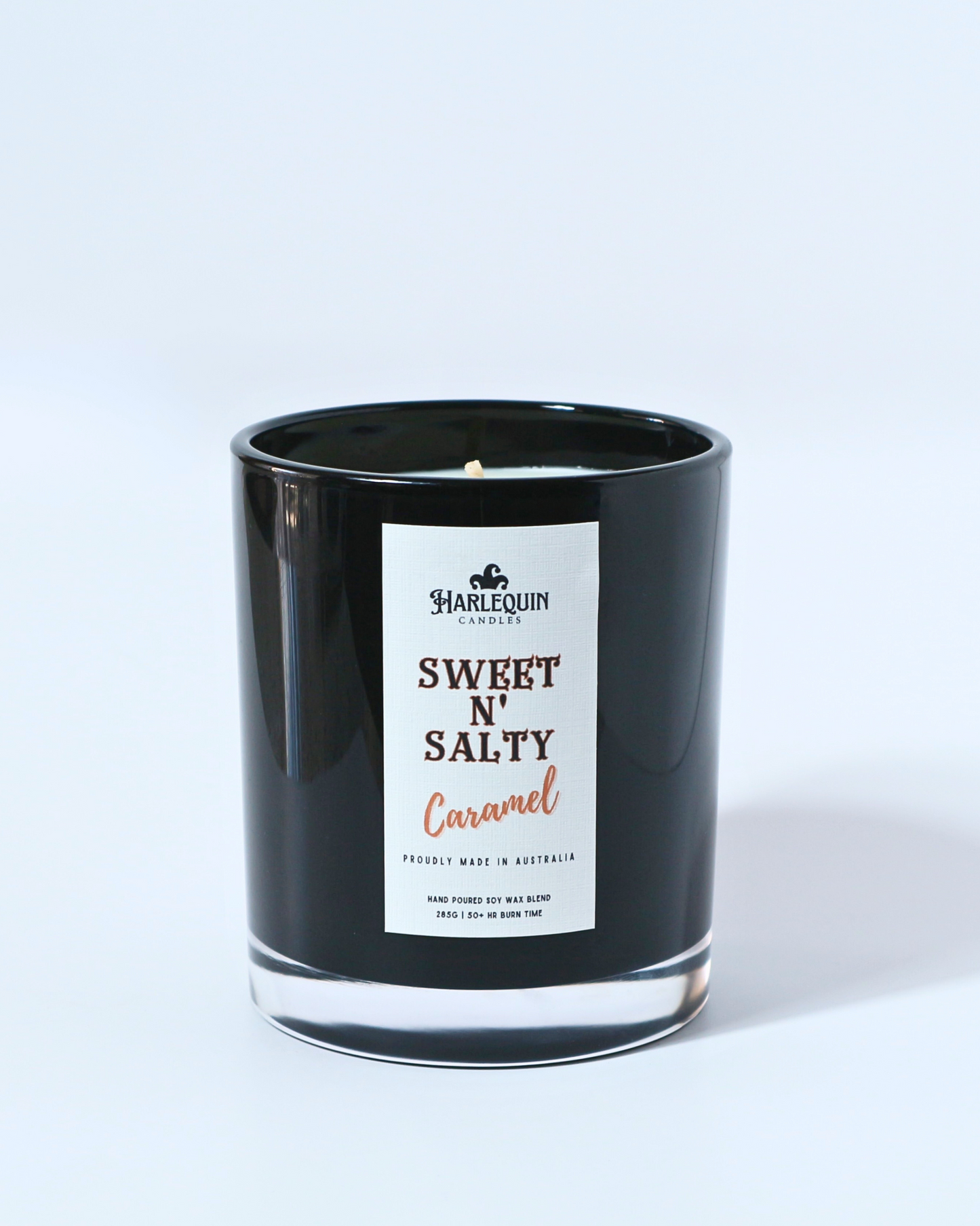 A Harlequin Candles "Sweet N' Salty Caramel" hand-poured soy wax candle in a black glass jar, styled beside a white vase with flowers and stacked books. Premium Australian made scented candle.