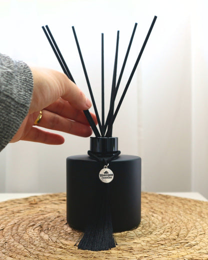 Close-up of a hand adjusting black reeds within a matte black Harlequin Candles reed diffuser bottle, adorned with a black tassel and silver logo charm. Demonstrates the easy activation process for your home fragrance diffuser on a woven mat.