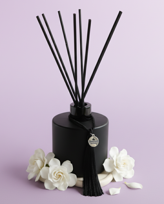 An Australian made Harlequin Candles reed diffuser featuring gardenia and lilac fragrance, perfect for a long-lasting floral home scent. The bottle is set against a purple background with white gardenia flowers.