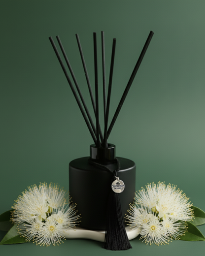 An Australian made Harlequin Candles reed diffuser with a fresh floral home fragrance of orange blossom and lemon myrtle, set against a vibrant green background with Australian Lemon Myrtle blossoms and green leaves.