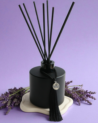An Australian made Harlequin Candles black matte reed diffuser with a calming and relaxing fragrance of lavender and vanilla, set against a purple background with lavender sprigs.