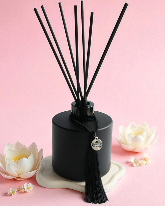 An Australian made Harlequin Candles black matte reed diffuser featuring a delicate floral home fragrance of white lotus, placed on a pink background with jasmine flowers.