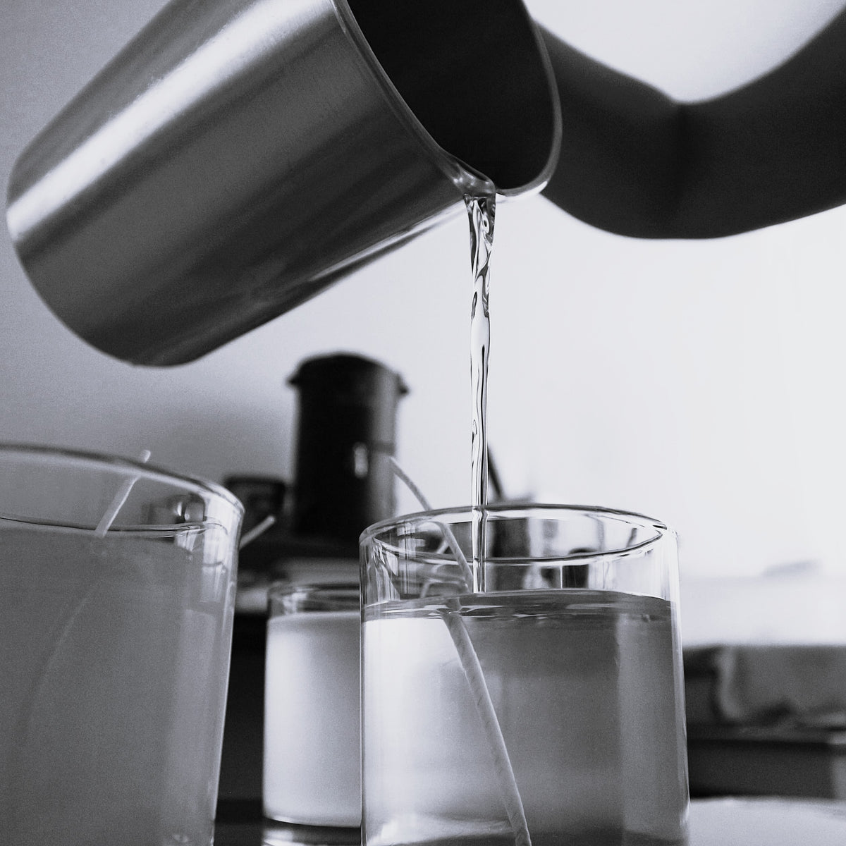 Monochrome close-up of melted soy wax being poured from a metal pitcher into a clear glass candle jar with a centered wick, illustrating the meticulous process of handcrafting Harlequin Candles soy candles.