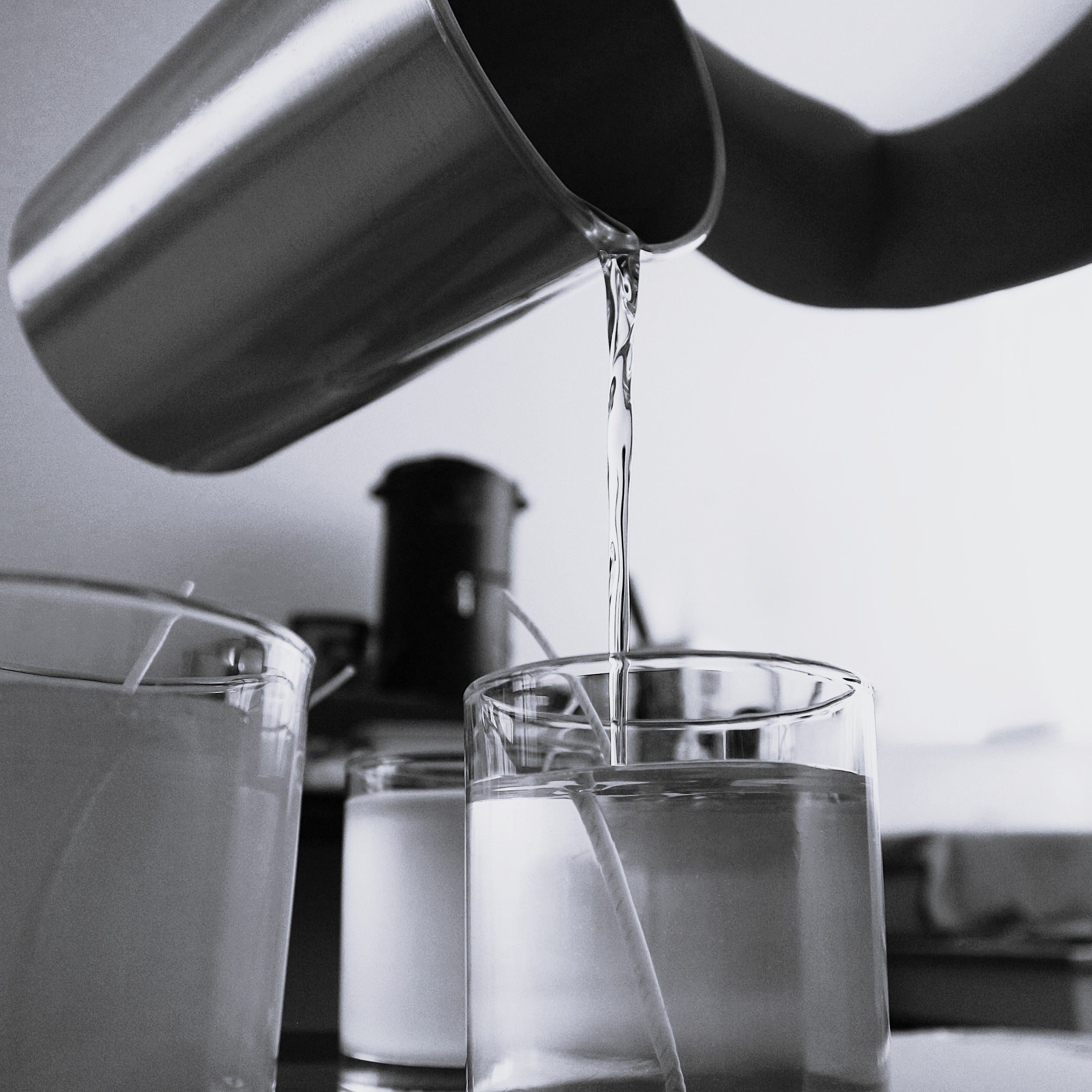 Monochrome close-up of melted soy wax being poured from a metal pitcher into a clear glass candle jar with a centered wick, illustrating the meticulous process of handcrafting Harlequin Candles soy candles.