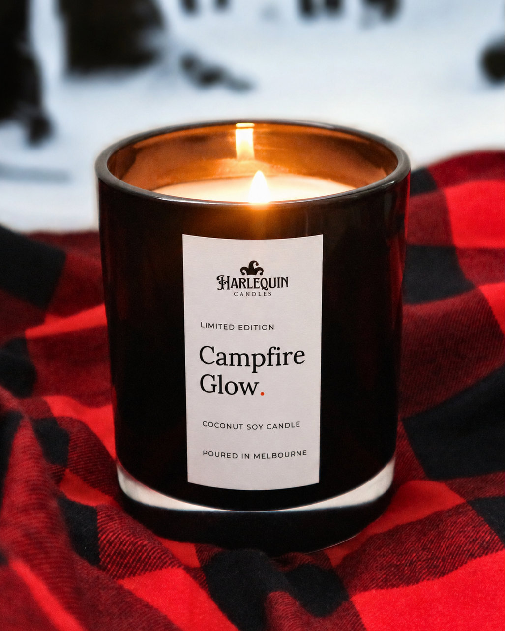 Campfire Glow smoky wood coconut soy candle burning in a black glass jar on a red checked blanket, hand-poured in Melbourne for a cosy winter fireside scent