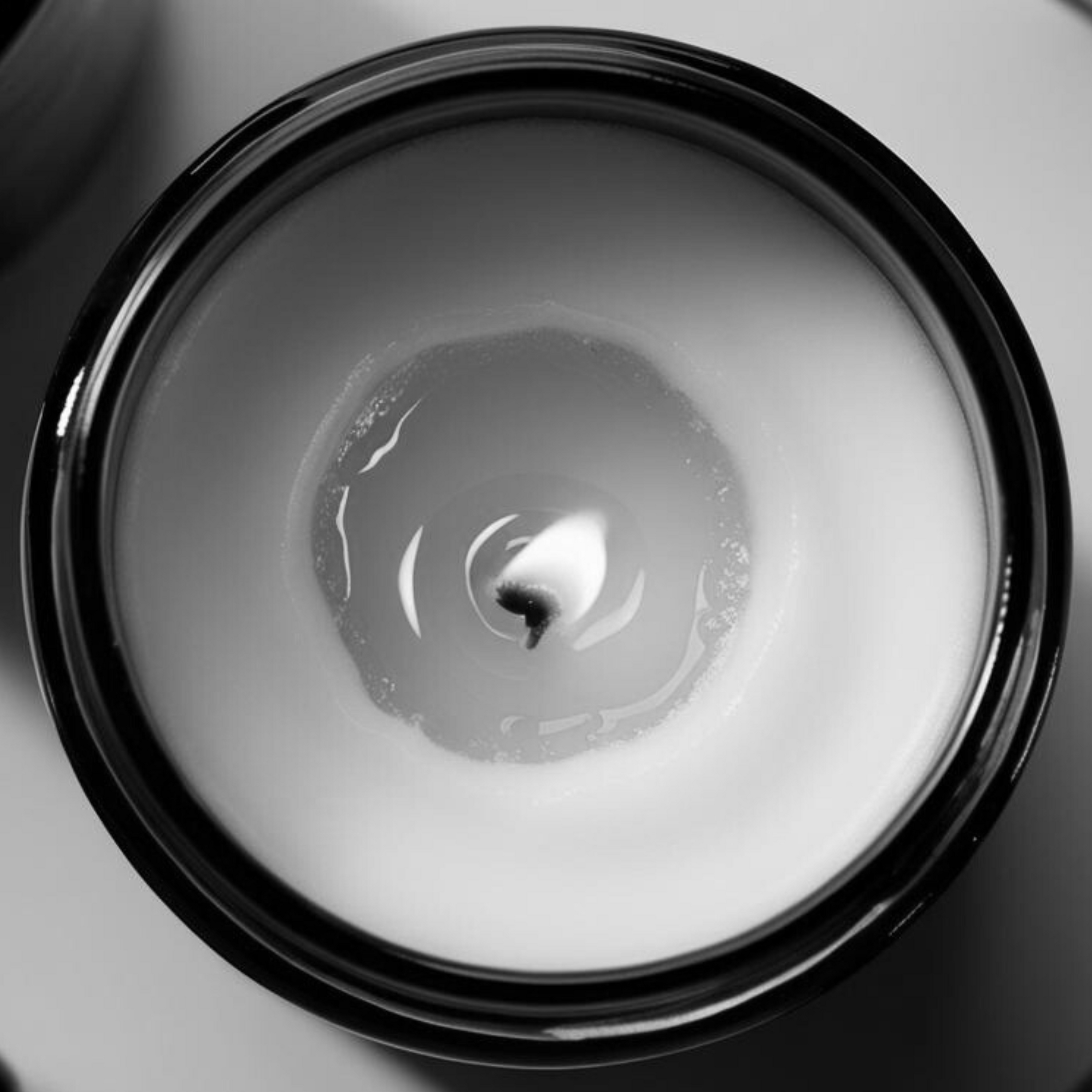 Monochrome overhead view of a lit Harlequin Candles soy candle in a black glass jar, clearly showing 'tunneling' where the wax has only melted down the center, leaving unmelted wax around the edges. Illustrates improper candle burning for a candle care guide on preventing tunneling.
