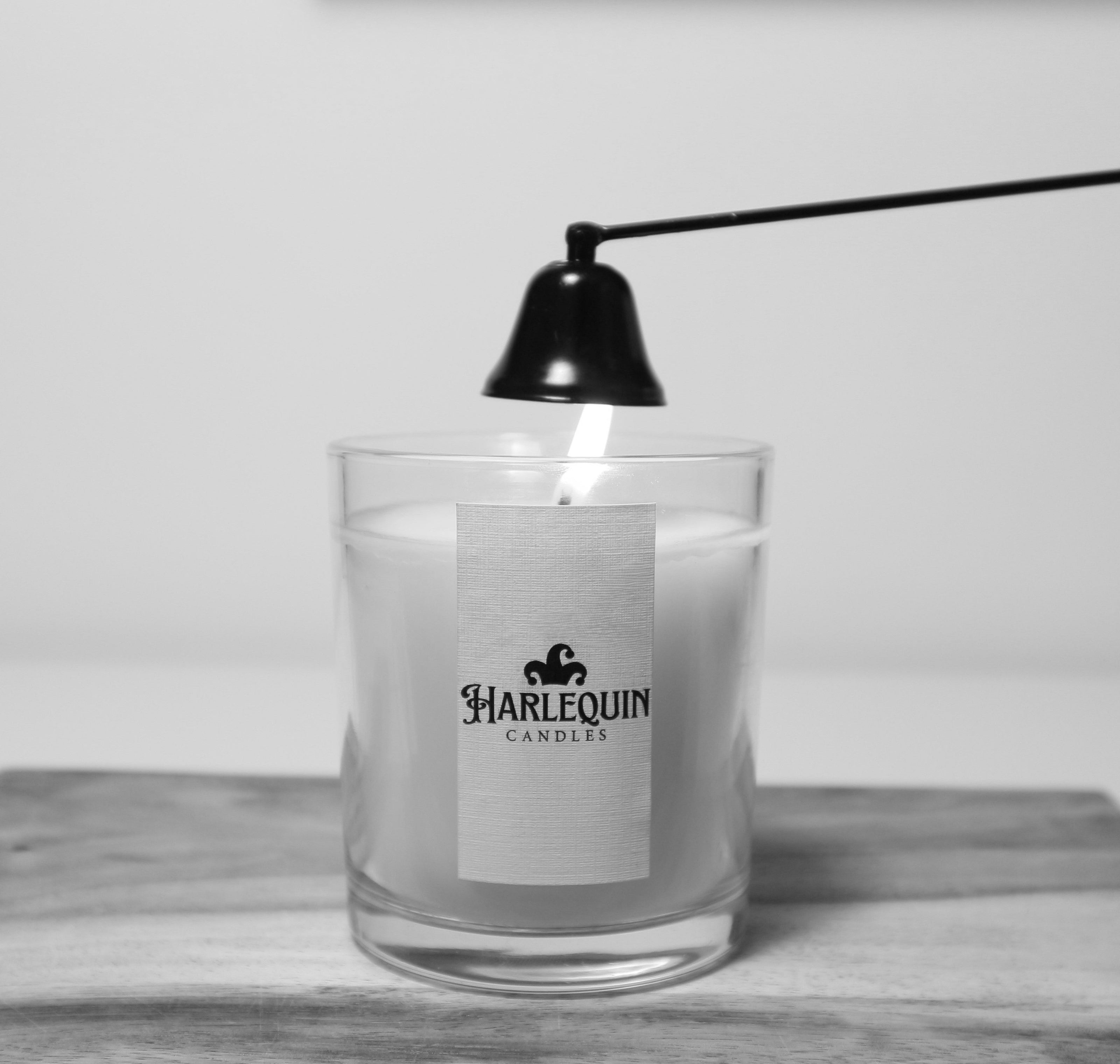 A black candle snuffer is used to safely extinguish the flame of a lit Harlequin Candles soy candle in a clear glass jar, resting on a wooden board. Demonstrates essential candle care for preventing smoke and ensuring safety.