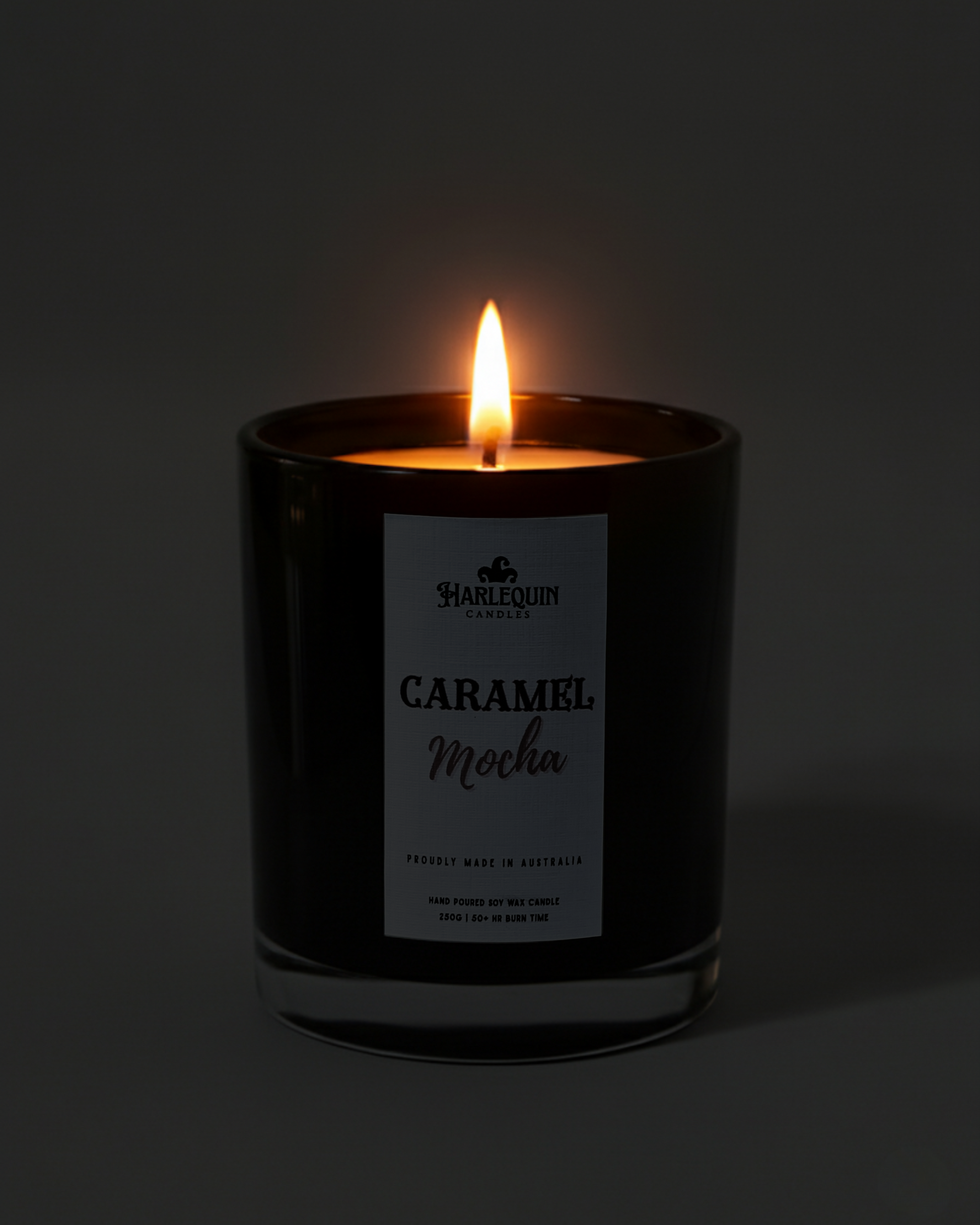 Lit Harlequin Candles Caramel Mocha candle in a dark glass jar against a dark background, emphasizing the warm, cozy glow of the flame. Describes a proudly Australian-made, hand-poured soy wax candle.