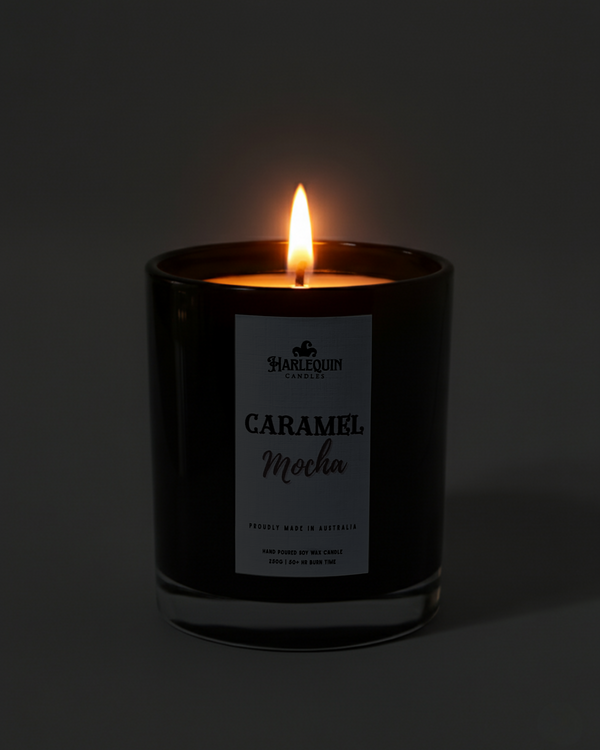 Lit Harlequin Candles Caramel Mocha candle in a dark glass jar against a dark background, emphasizing the warm, cozy glow of the flame. Describes a proudly Australian-made, hand-poured soy wax candle.