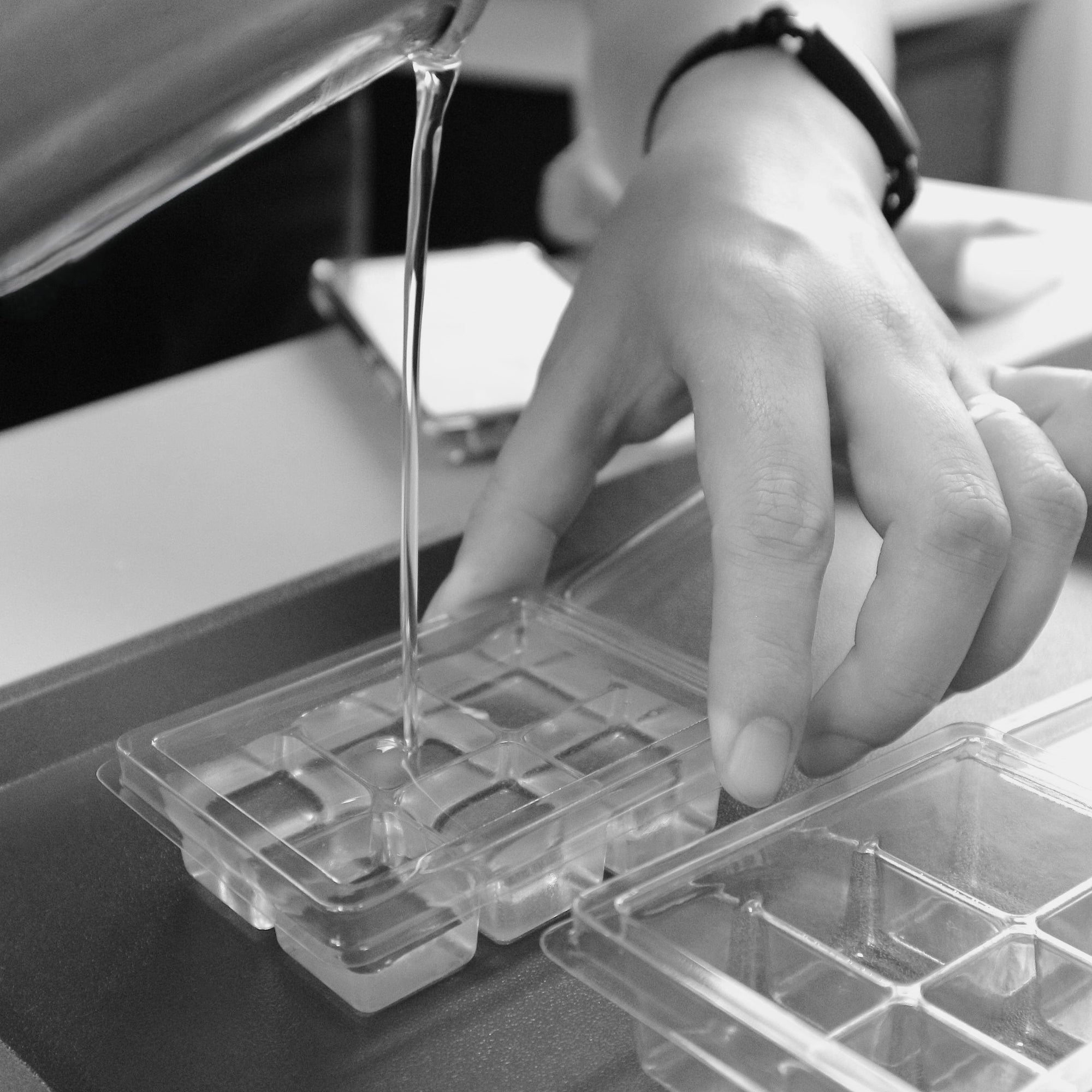 Monochrome close-up of liquid soy wax being meticulously poured into clear plastic molds, held by a hand, illustrating the artisan process of crafting Harlequin Candles wax melts.